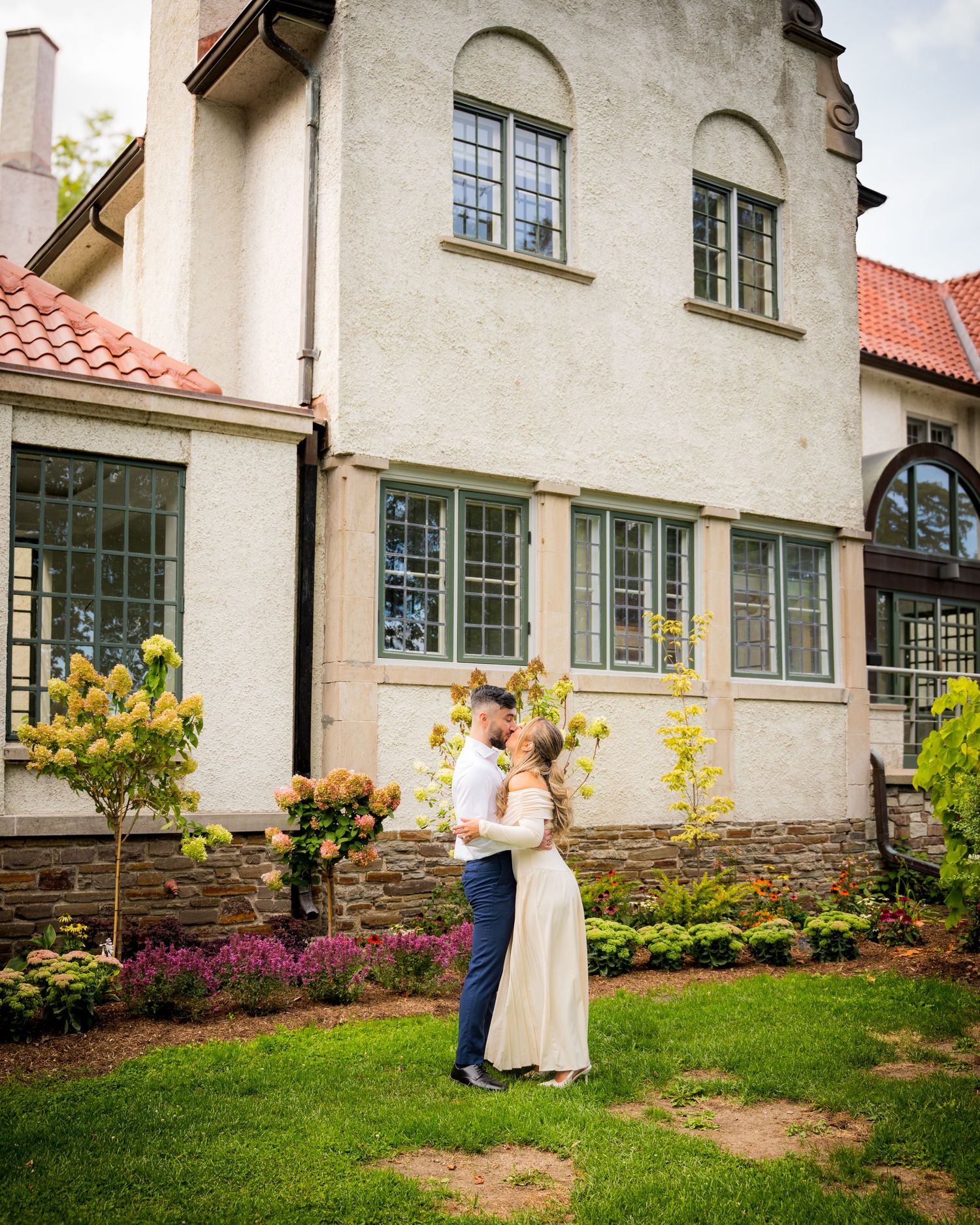 A couple shares a romantic moment on the lawn of a beautiful historic building with stone and stucco exterior.