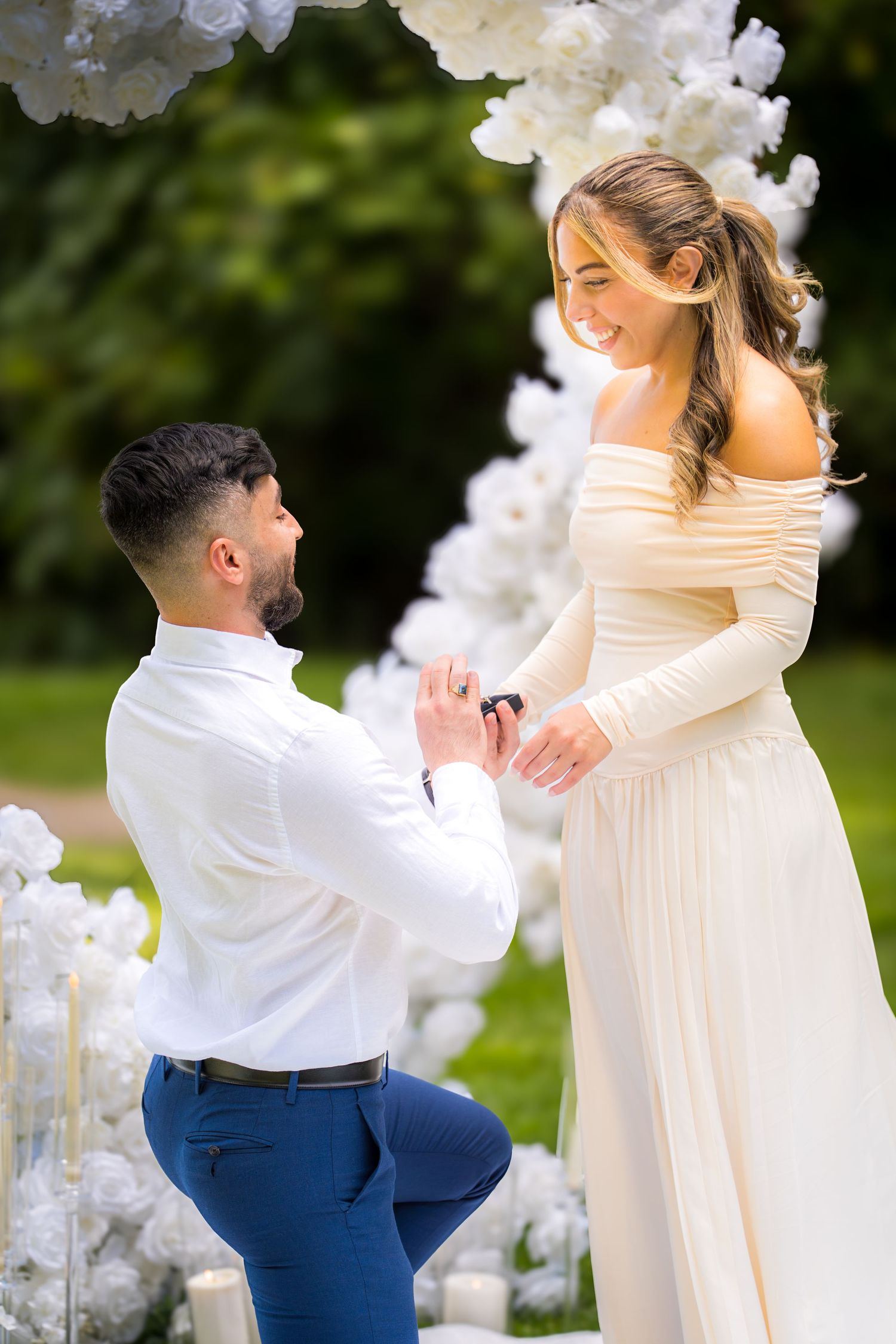 A romantic proposal scene in a garden with white floral decorations and a couple in formal attire.