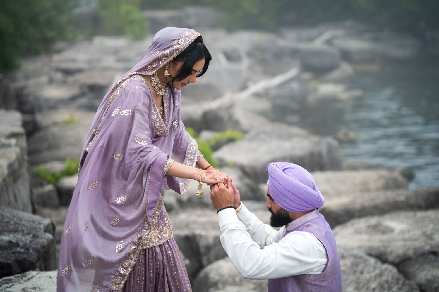 Couple in lavender wedding attire pose together on rocky terrain near water.