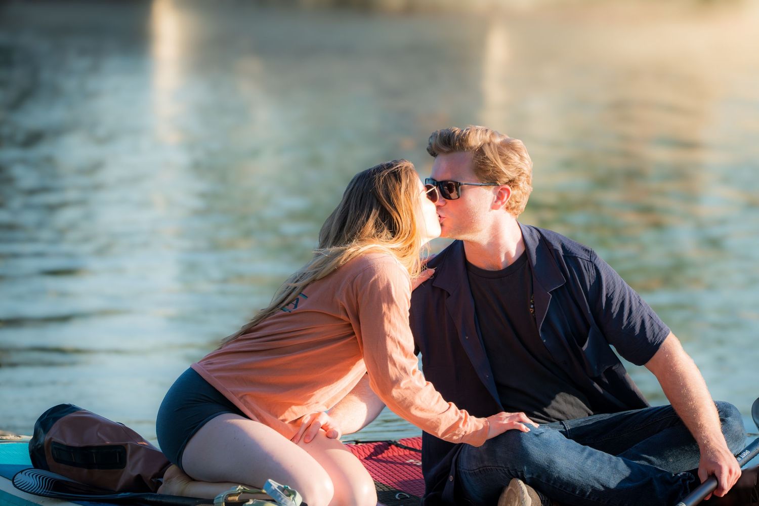 A couple sharing a romantic kiss while sitting on a paddleboard on a calm lake at sunset.