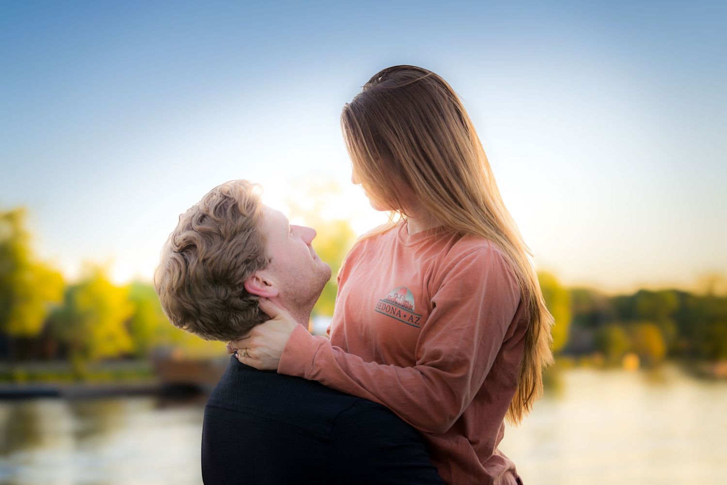 Romantic couple embracing and gazing at each other by a lake during a beautiful golden sunset.