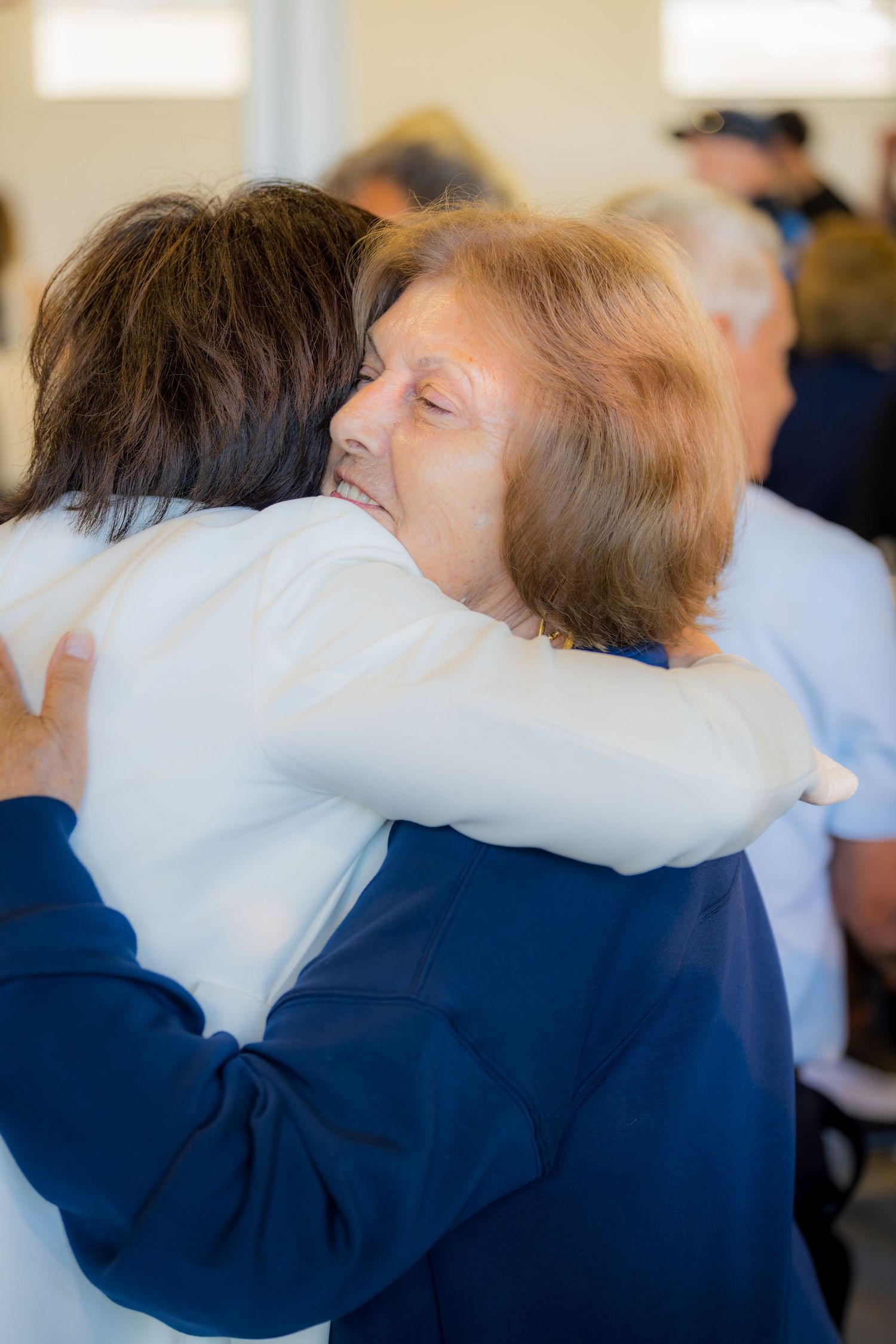 Two women share a warm embrace at a social gathering, one smiling with eyes closed.