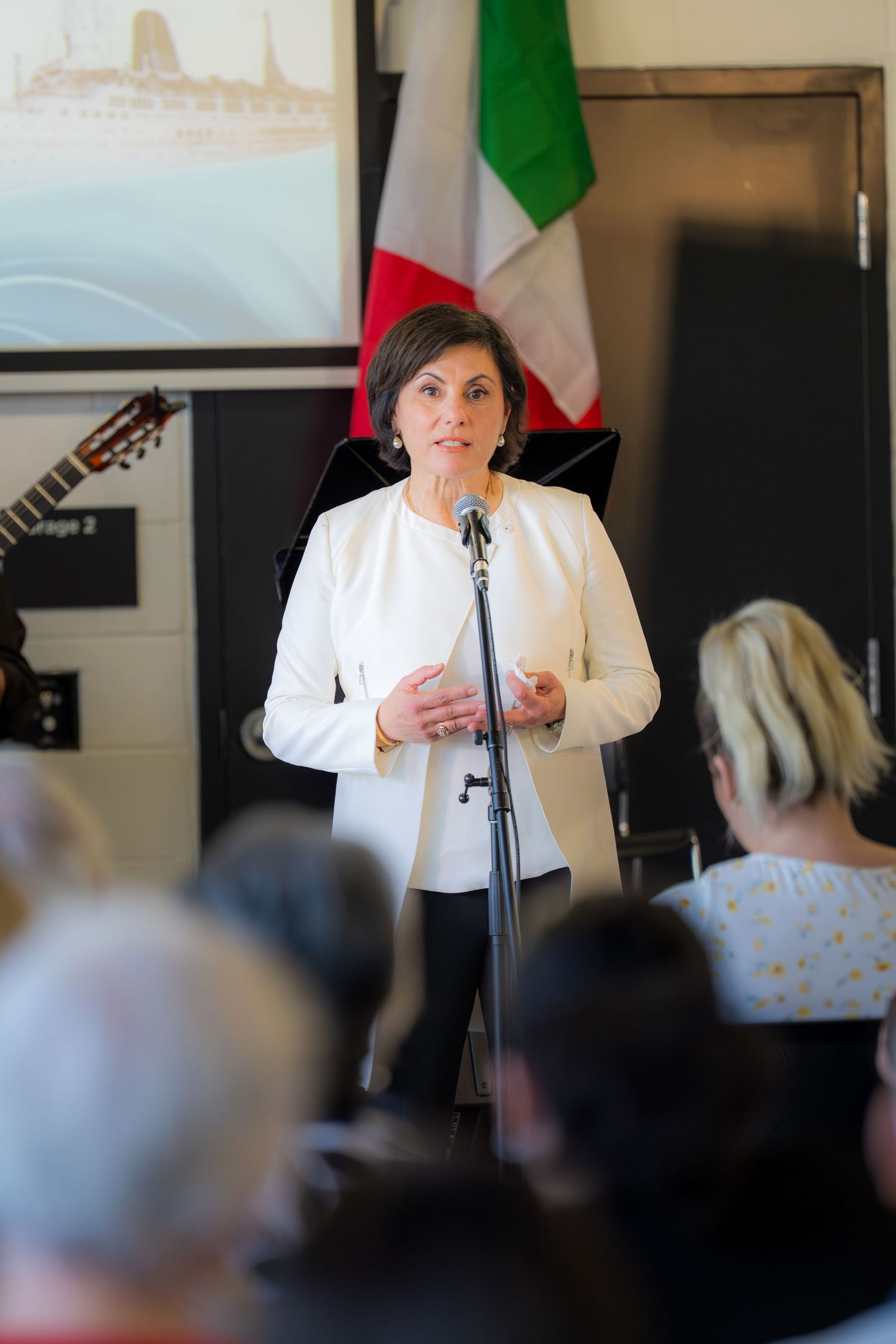 Woman in white blazer speaks at microphone podium before audience with Italian flag backdrop.