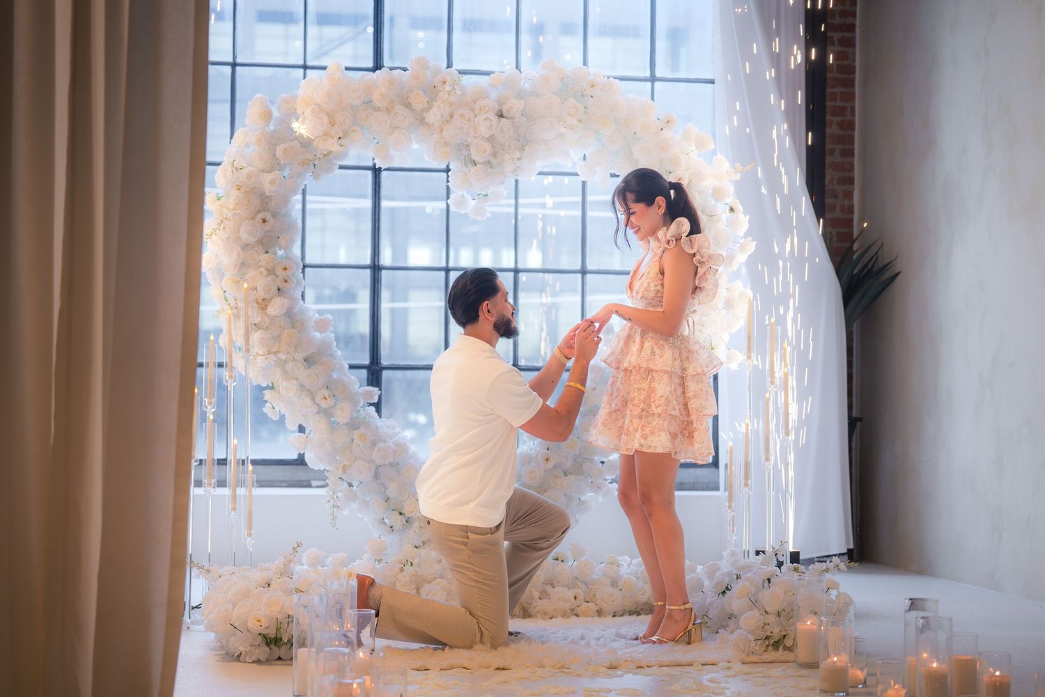 Man proposing on one knee to his partner under a white floral heart arch with candles and flowers.
