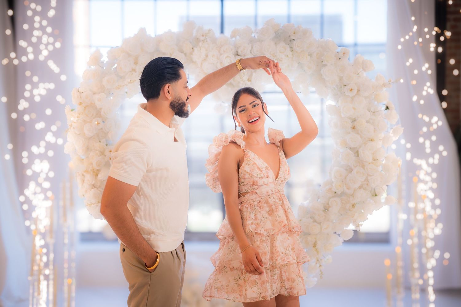 Joyful couple dancing under a white floral heart arch surrounded by twinkling fairy lights.