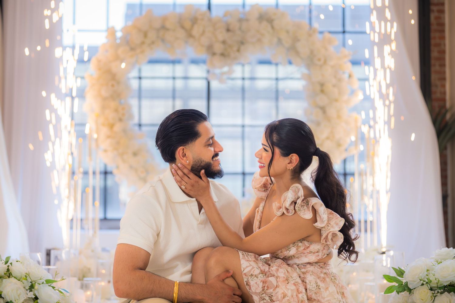 Couple sitting together gazing lovingly in front of a glowing white heart floral arch.