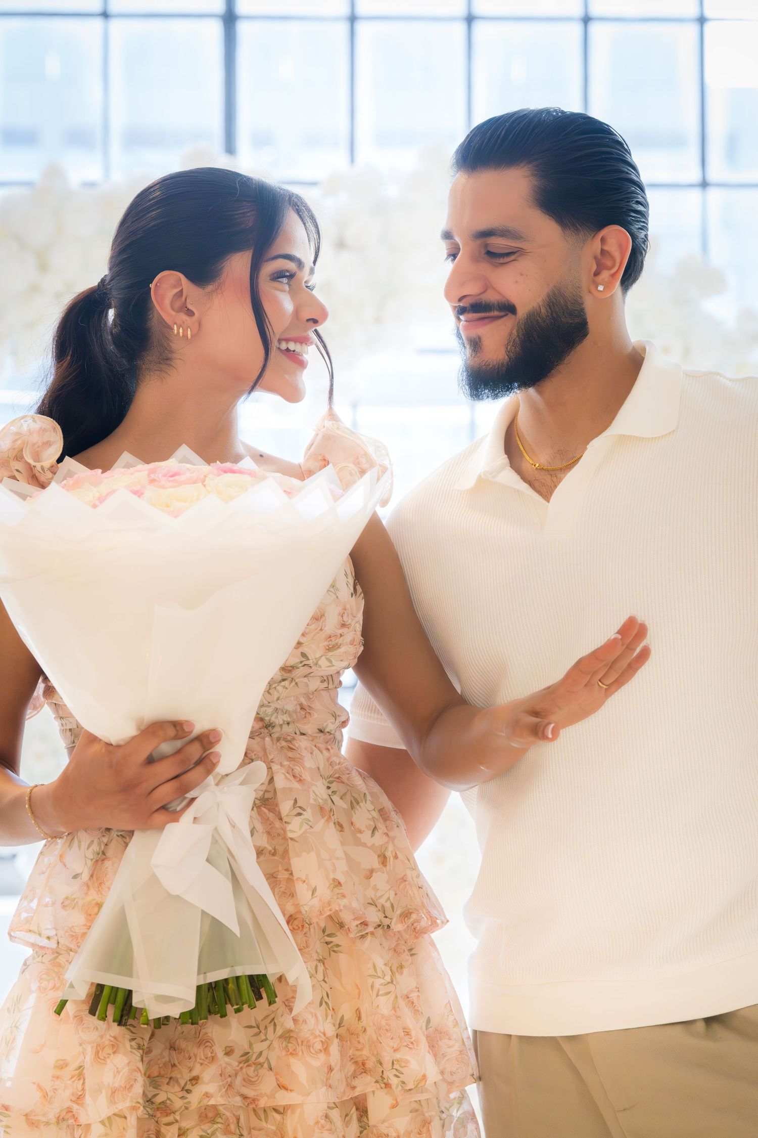 Smiling couple sharing a tender moment, woman holding white bouquet in elegant floral mini dress.