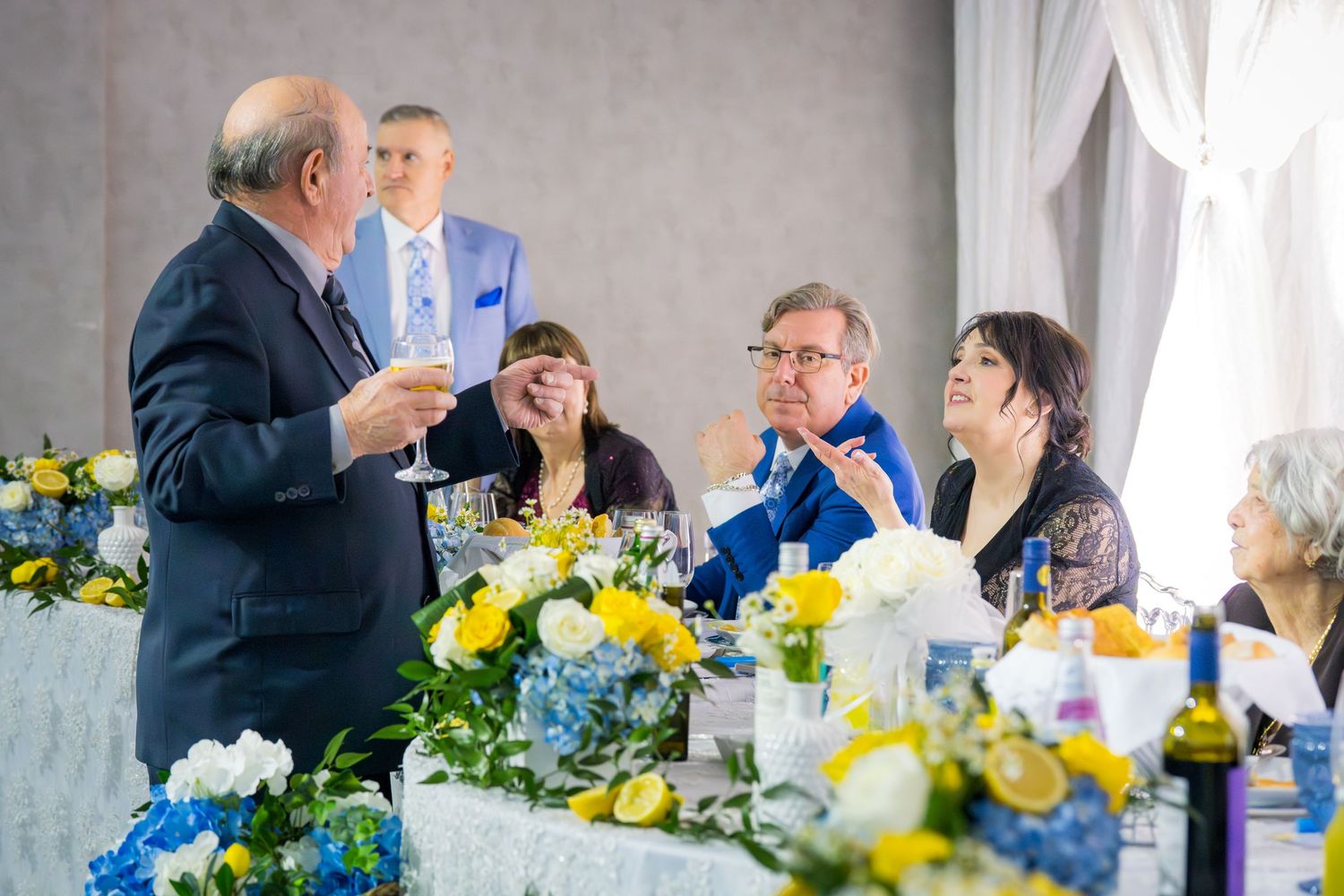 Man in suit making a toast with champagne at a wedding reception with blue and yellow floral centerpieces.