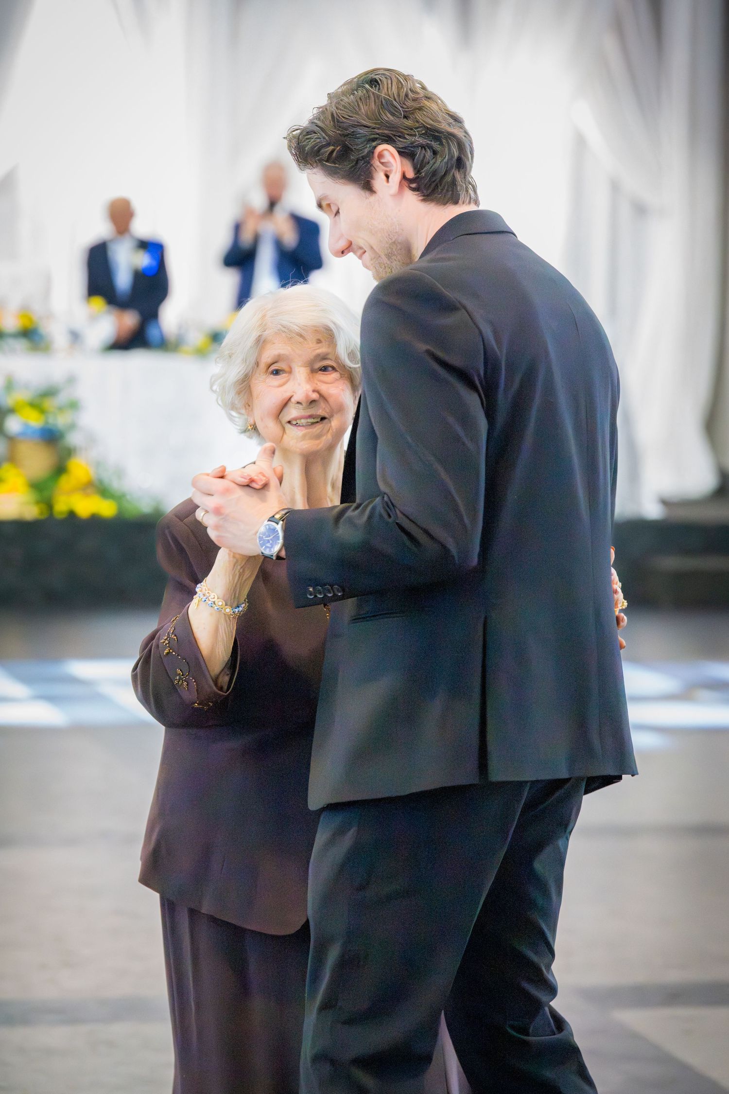 Young man in dark suit dancing with smiling elderly woman at a formal anniversary celebration event.