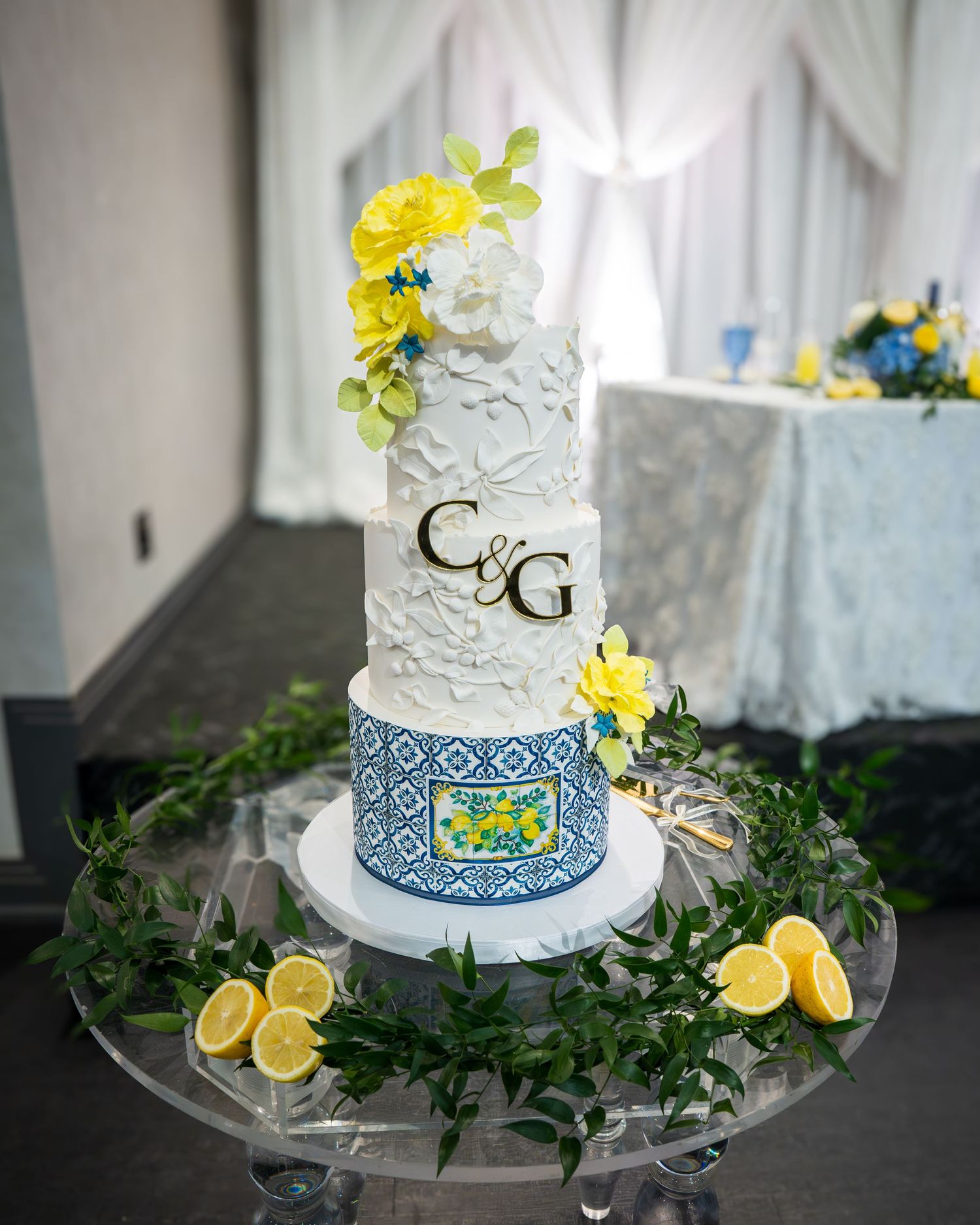 Three-tier white wedding cake with blue tile design, yellow flowers, and C&G monogram on a glass stand with lemons.