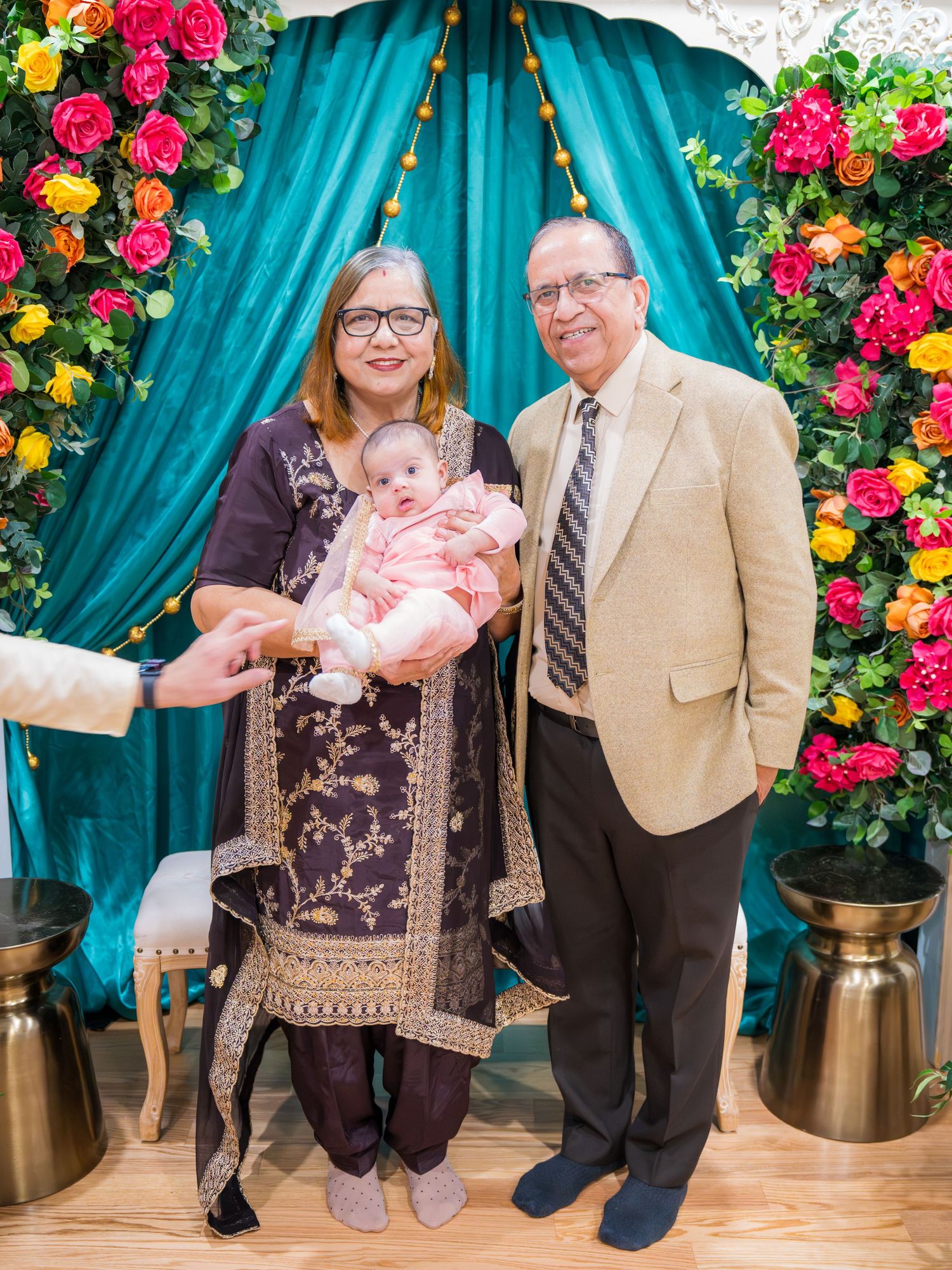 Couple in traditional South Asian attire holding a baby in front of a floral and teal drape backdrop.