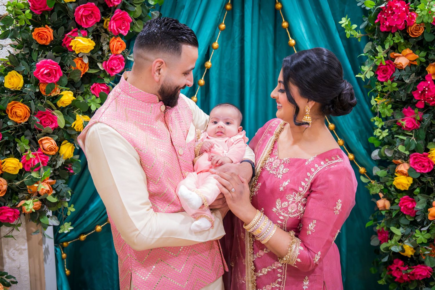 South Asian couple in pink traditional attire holding newborn baby in front of colorful floral backdrop.