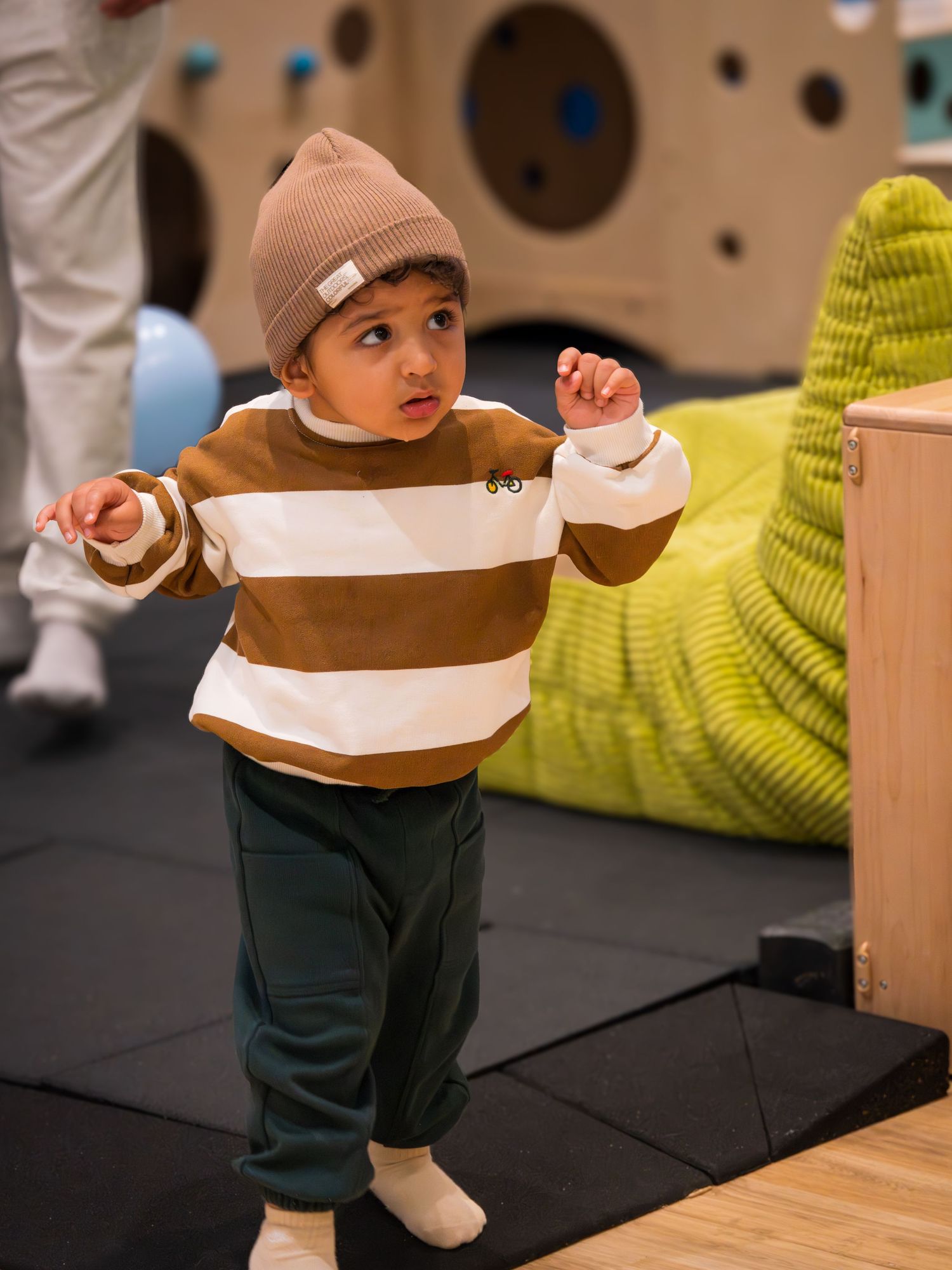 Toddler in brown striped sweater and beanie hat playing at an indoor playground.
