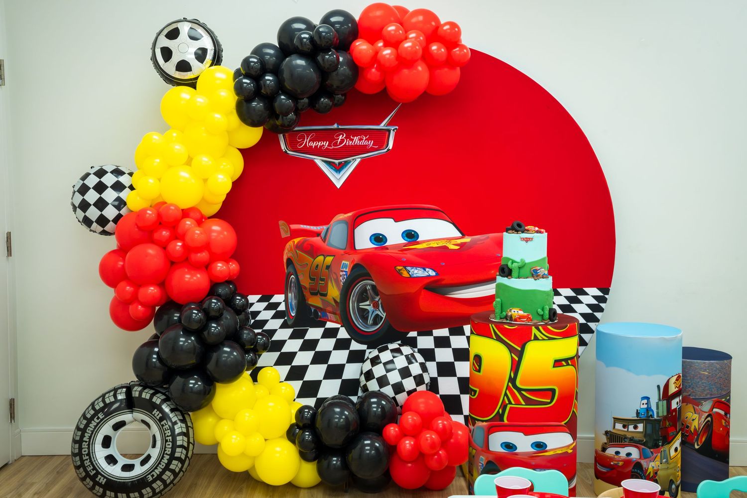 Cars-themed birthday party backdrop with Lightning McQueen, balloon garland in red, yellow, and black, and checkered flag decorations.