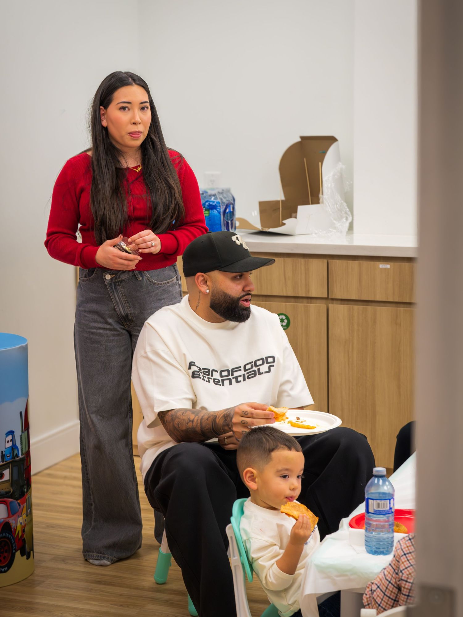 Woman in red sweater standing beside a man and toddler eating at a party.