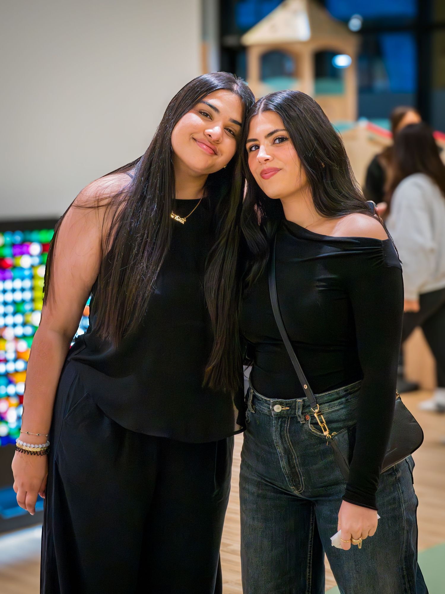 Two young women in black outfits pose together smiling at an indoor event.