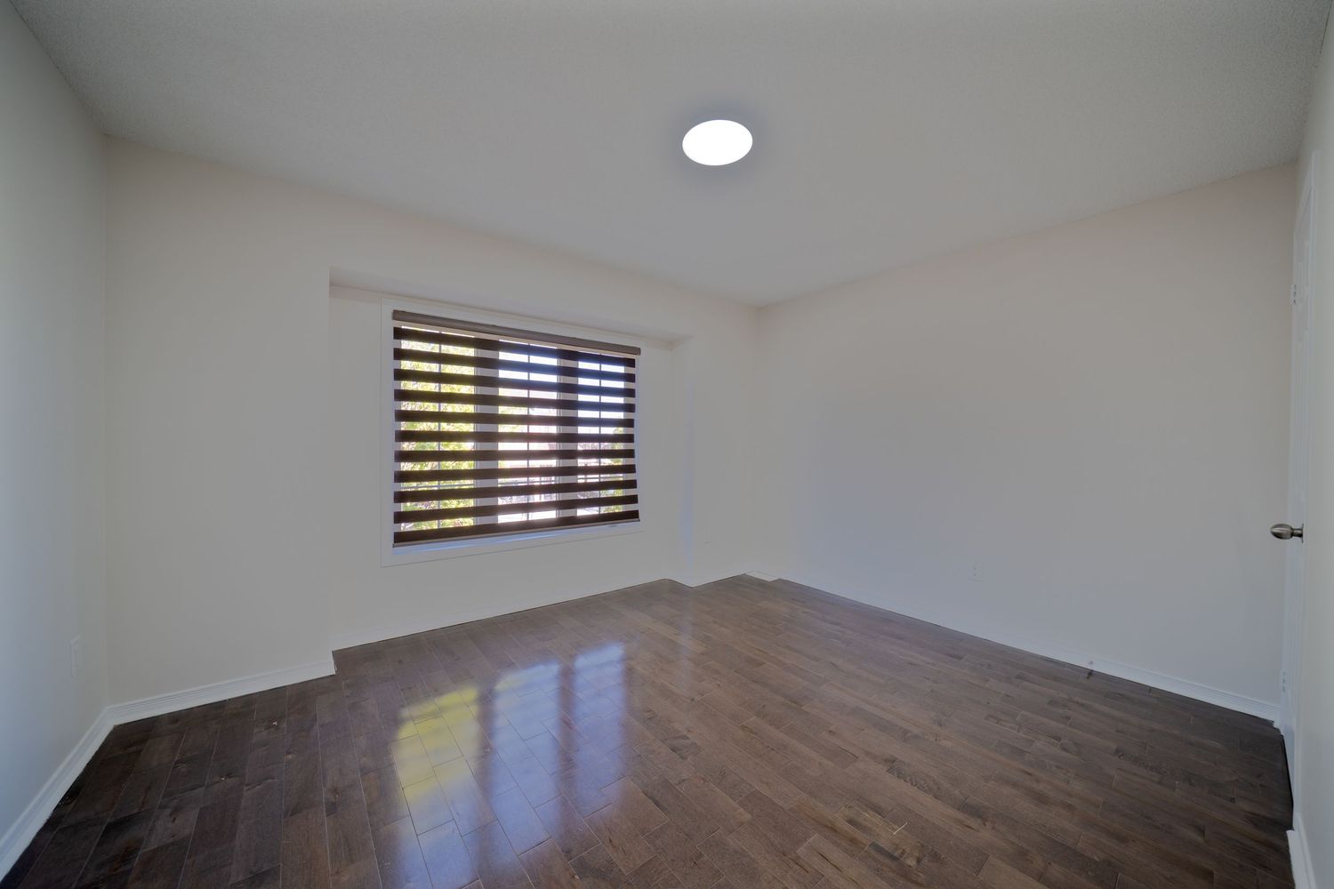 Empty room with hardwood flooring, white walls, and a window with horizontal blinds letting in natural light.