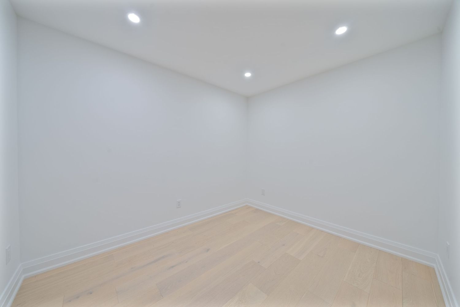 Empty room interior with light hardwood flooring, white walls, and recessed ceiling lights.