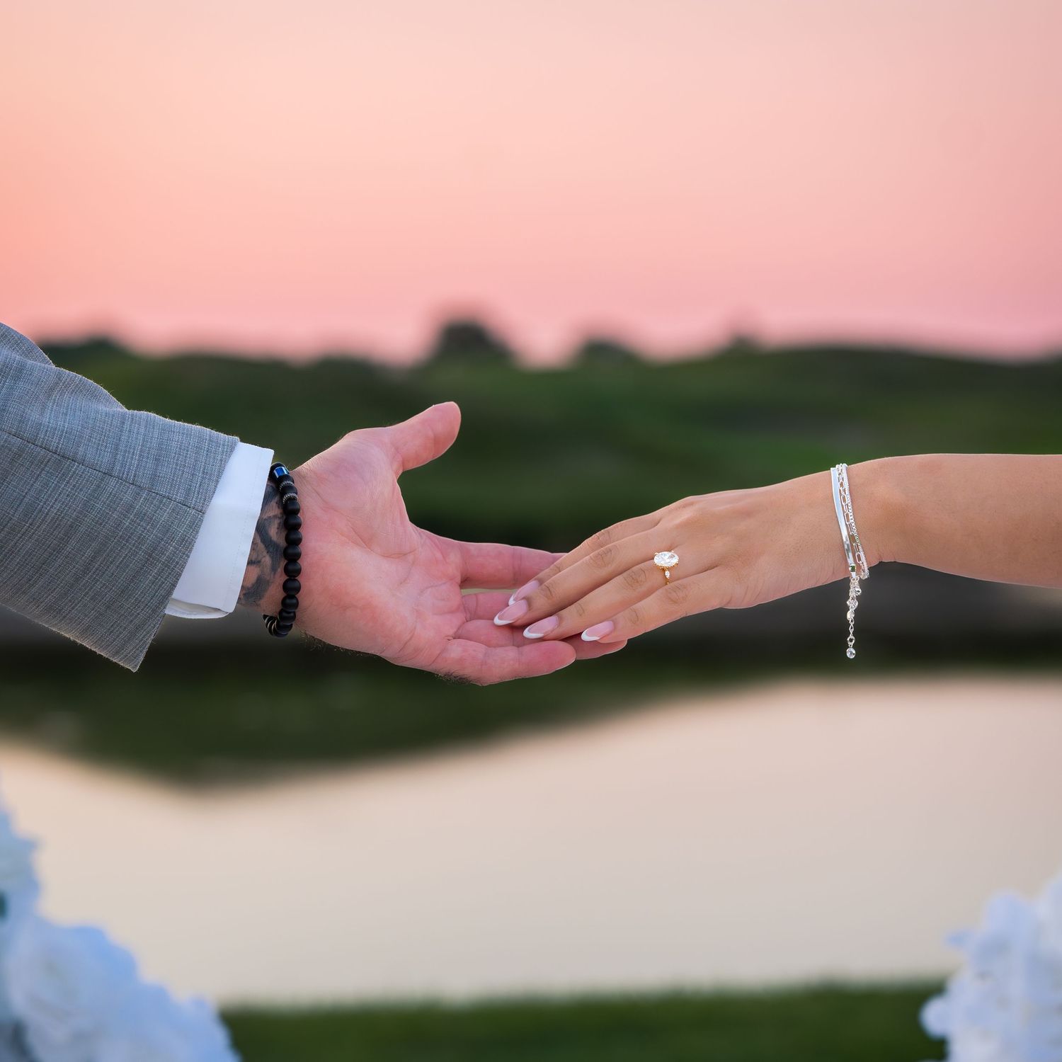 Hands reaching towards each other with engagement ring visible against sunset sky.