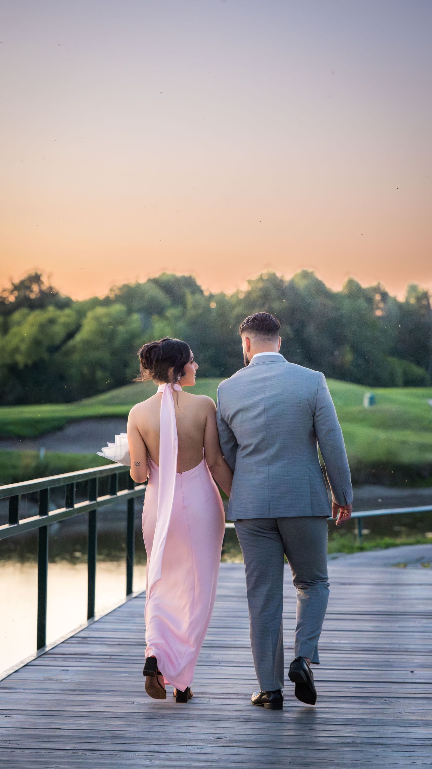 A romantic couple strolls along a waterfront boardwalk during golden hour.
