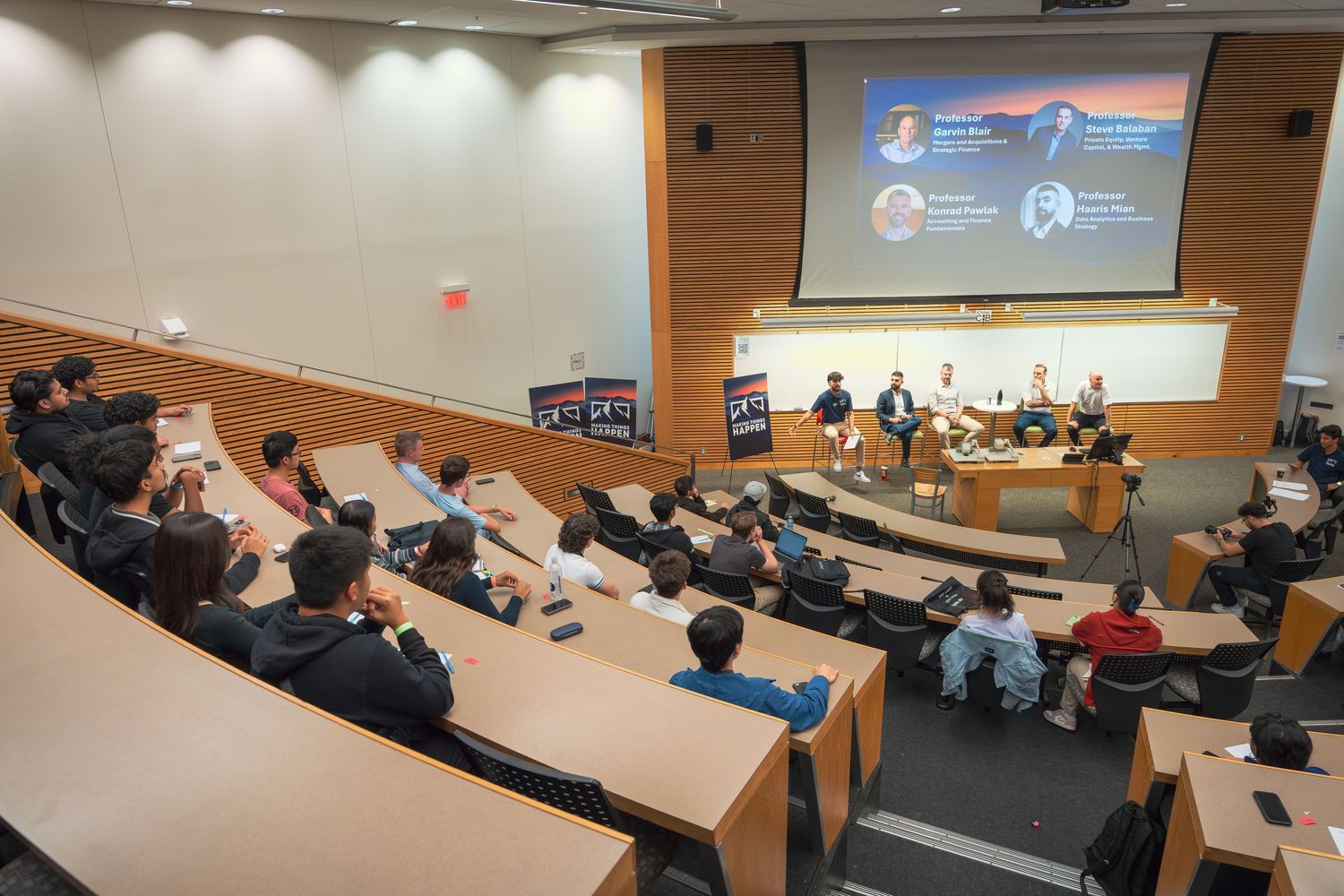 Students attend a lecture in a modern university classroom with tiered seating and projection screen.