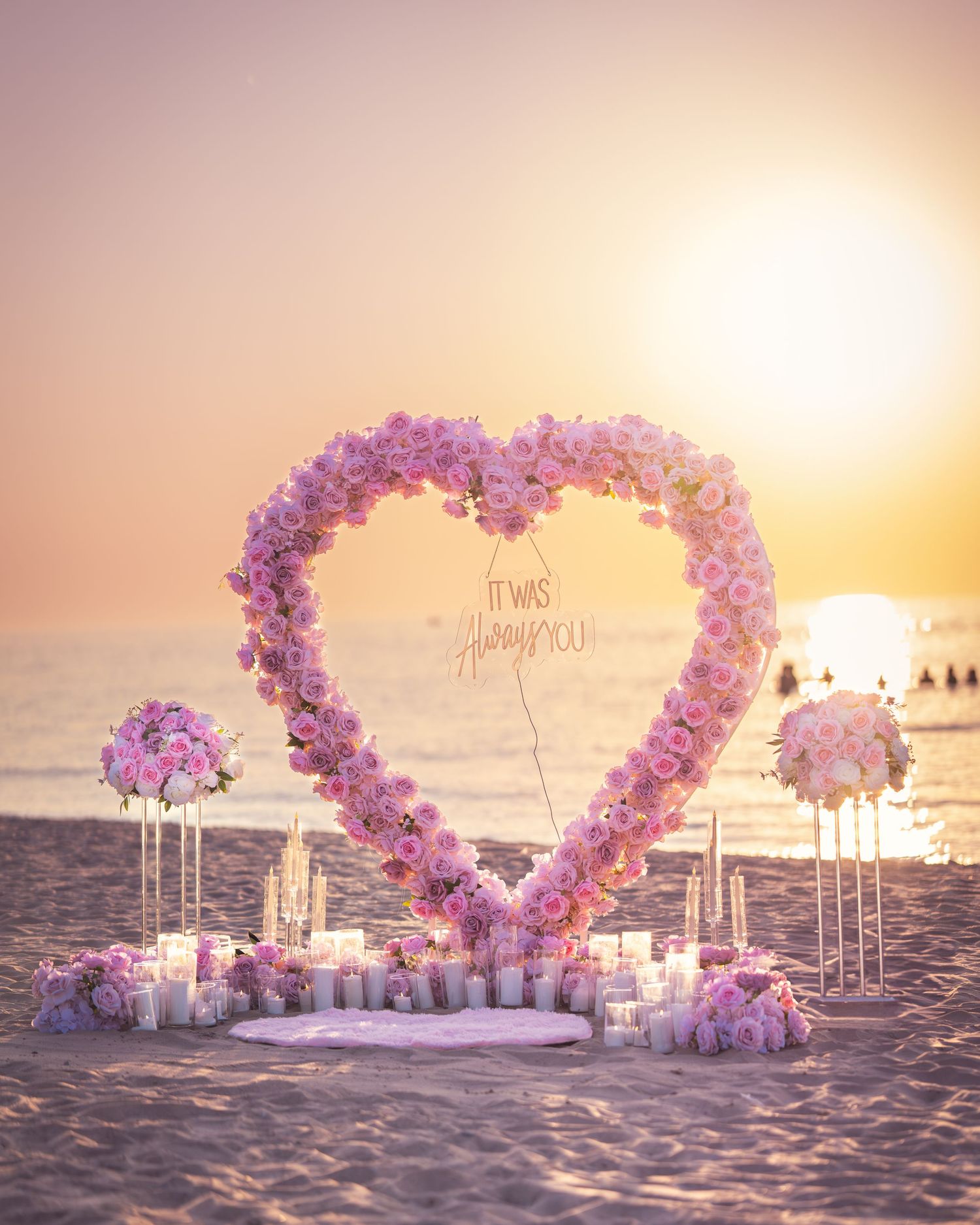Romantic heart-shaped floral arch with pink and purple flowers on a beach at sunset with candles and decor.