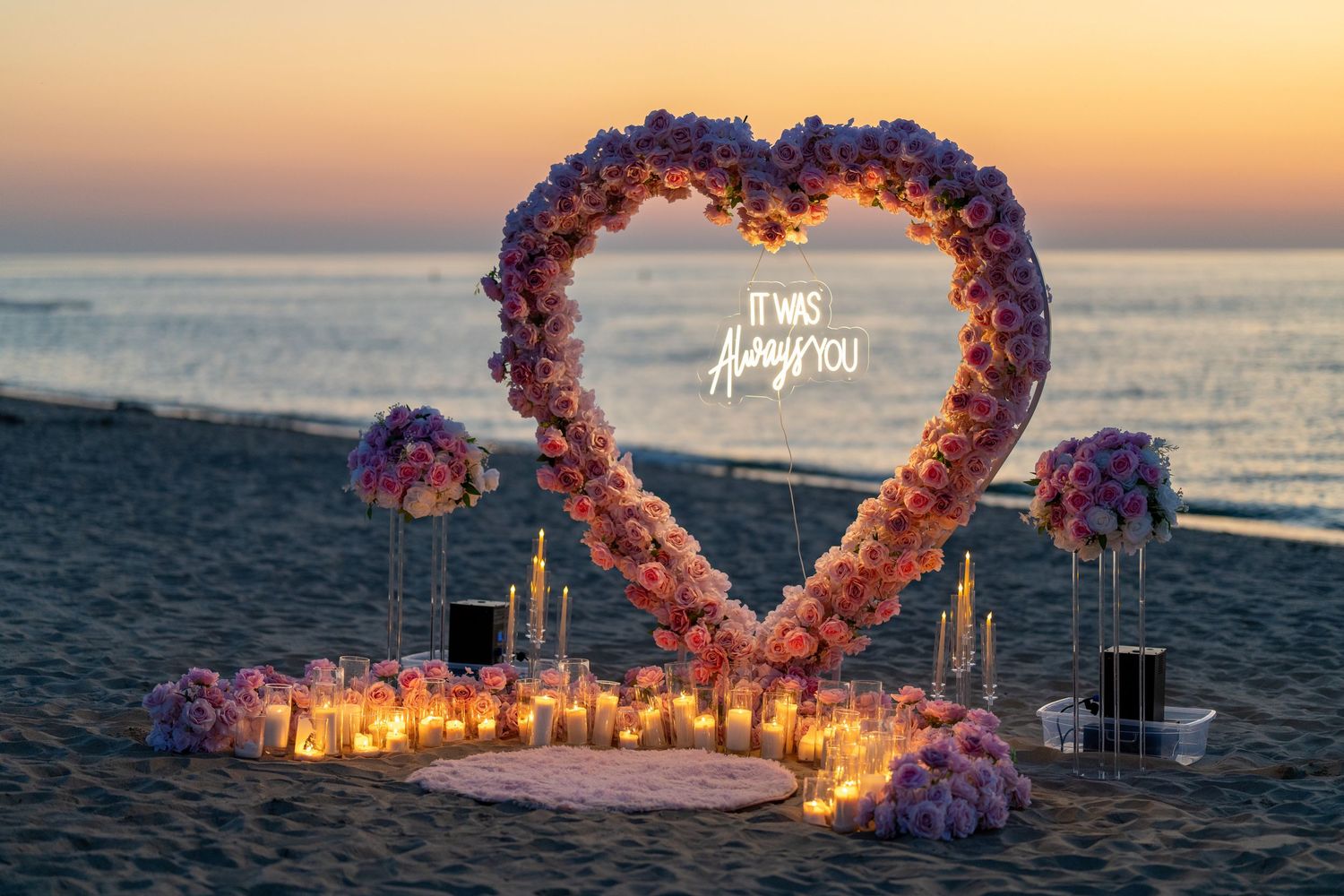 Sunset beach proposal setup with heart-shaped flower arch and glowing lanterns along the shore.
