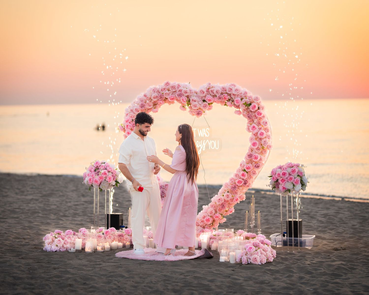 Elegant beach proposal setup with pink heart-shaped arch and lanterns at dusk.
