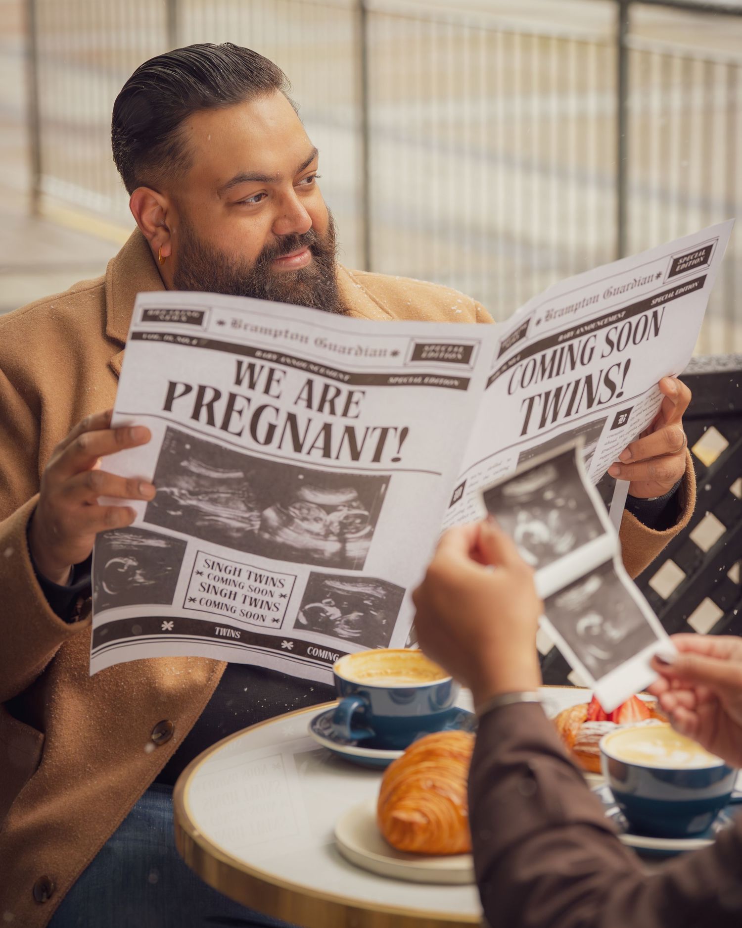 A person in a tan coat reads a custom newspaper with 'We Are Pregnant' headline at an outdoor cafe.