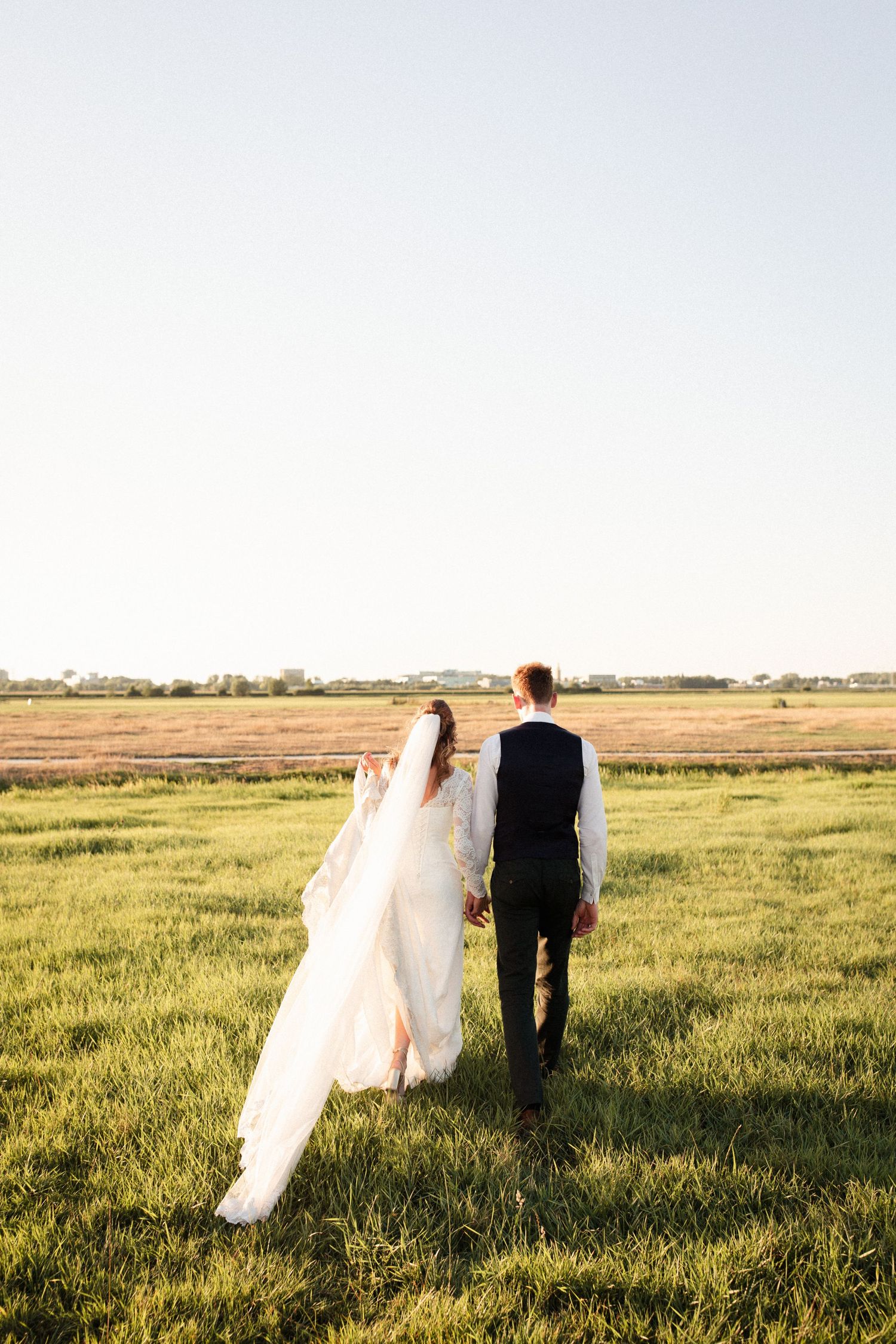 A couple in wedding attire walks through a sunlit meadow during golden hour.