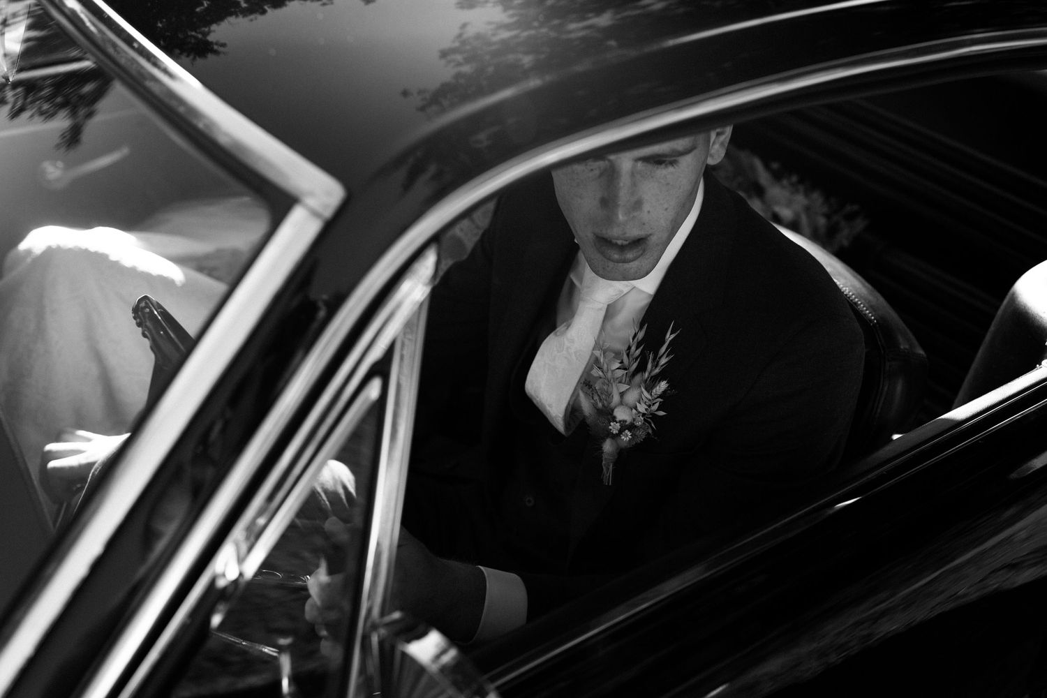 Black and white photo of someone wearing formal attire sitting inside a dark car.