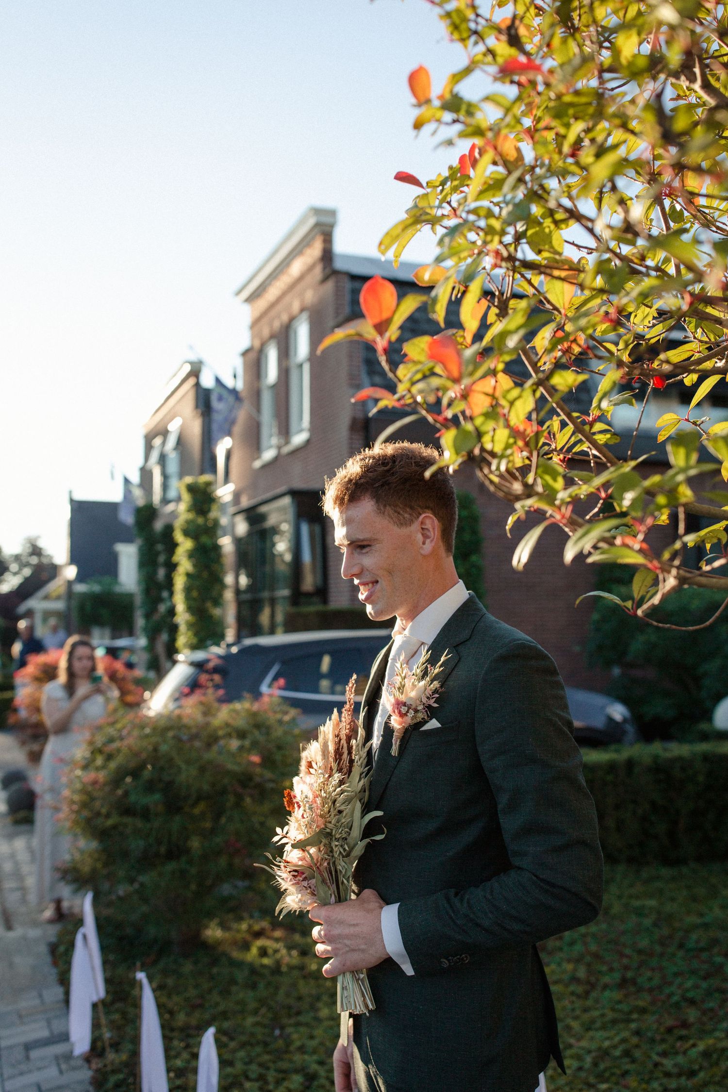 A groom in a black suit holds a delicate bouquet of flowers outside a brick building on a sunny autumn day.