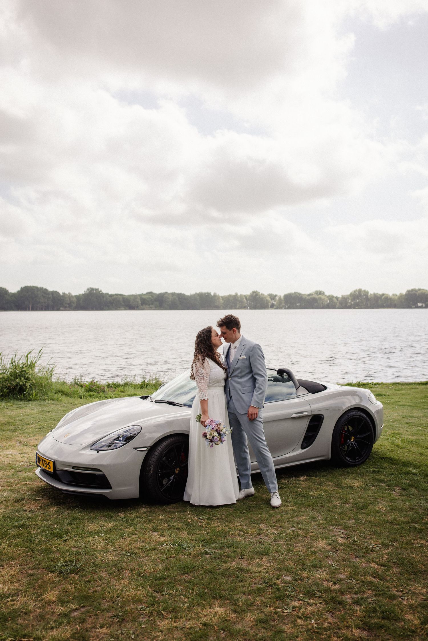 Elegant wedding photo featuring a silver luxury sports car on grass with water view.