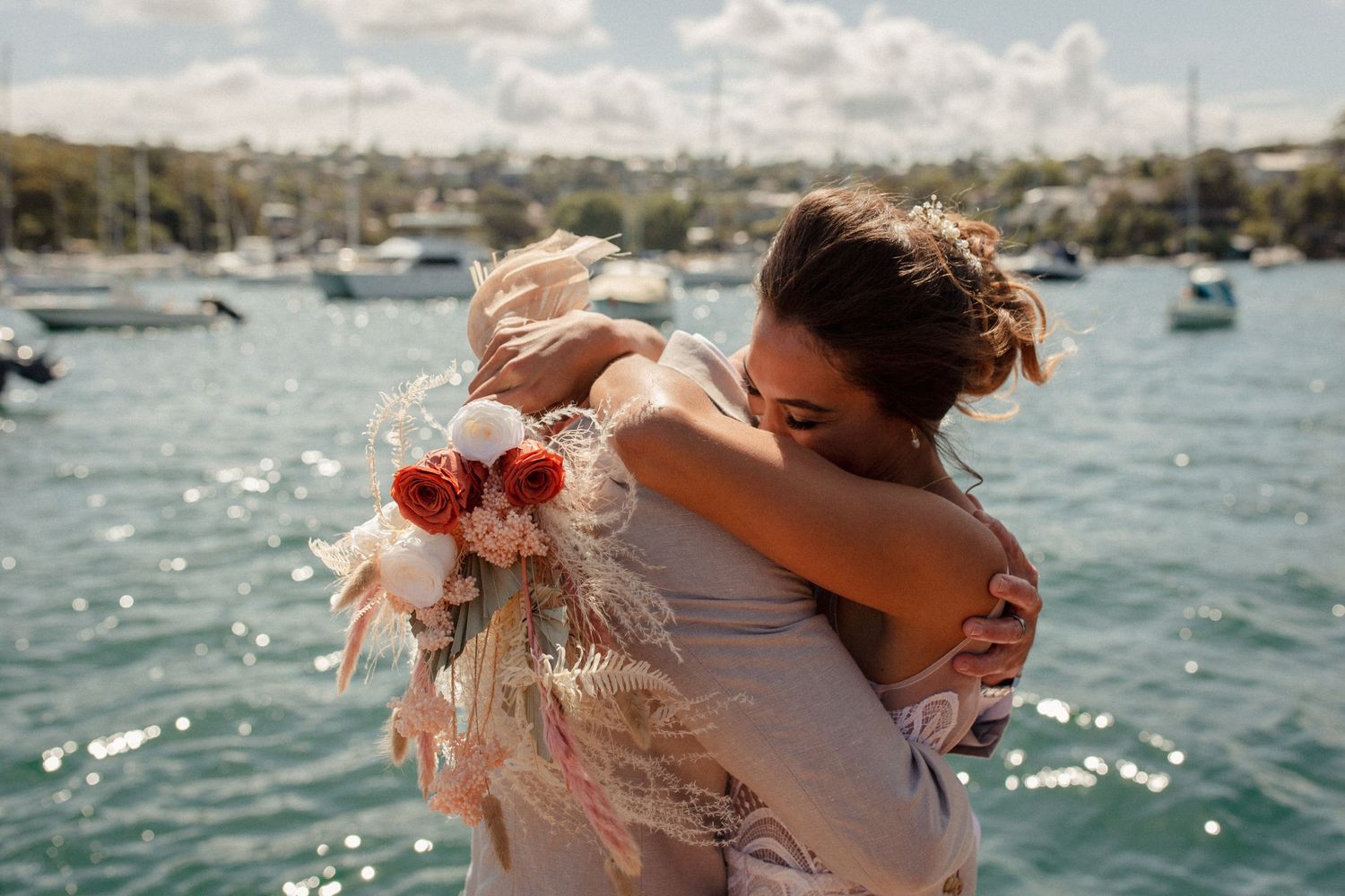 A joyful wedding embrace on a sunny dock with sparkling water in the background.