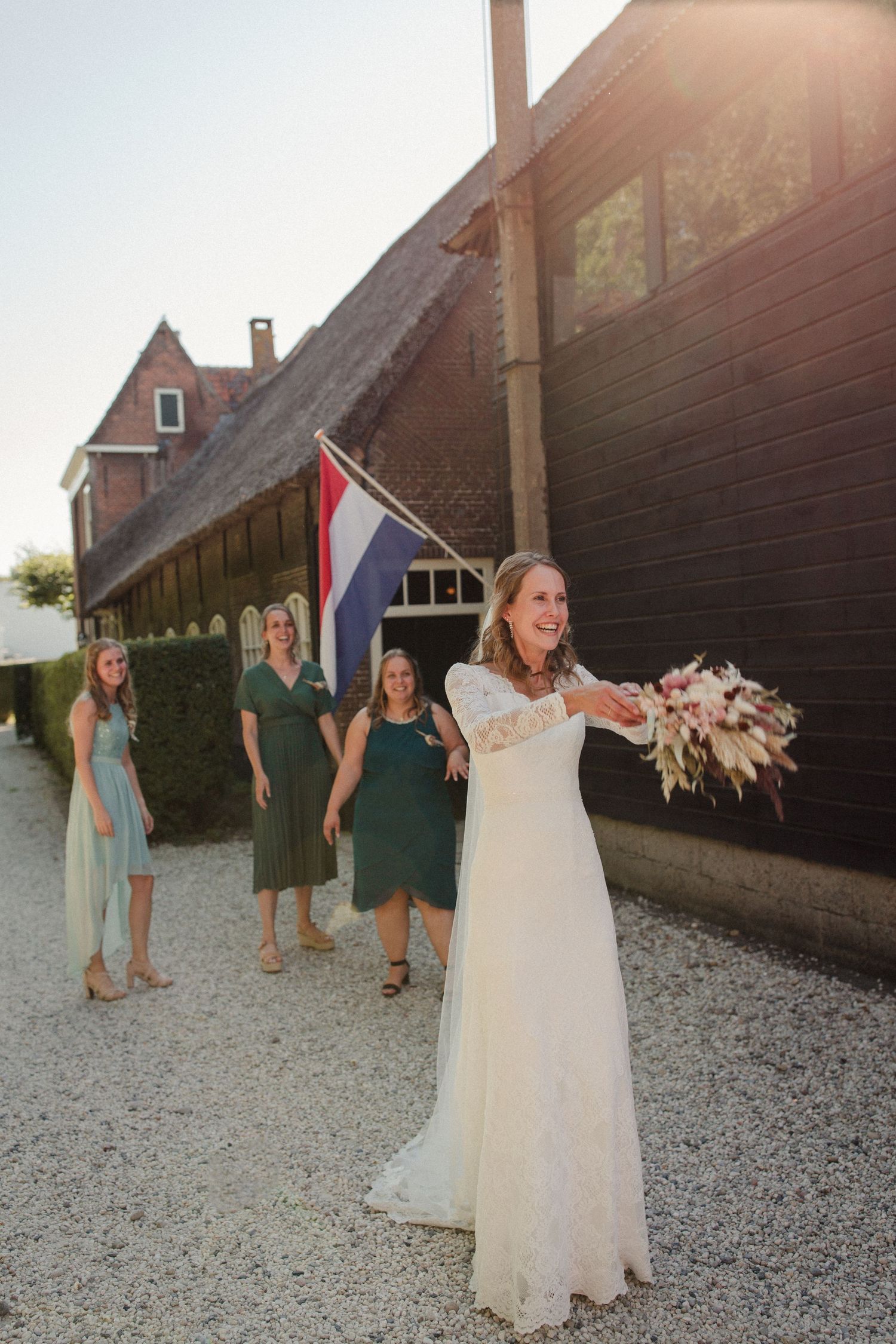 Wedding scene outside rustic barn with Dutch flag as bride prepares to toss bouquet at sunset.