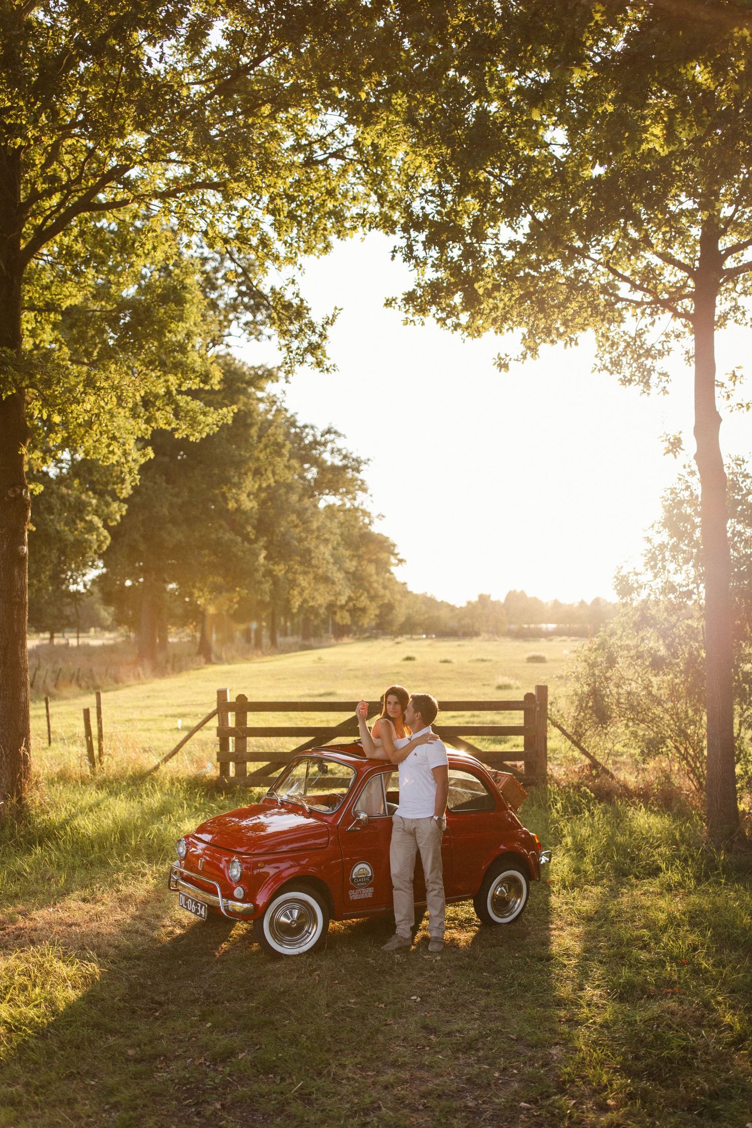 Koppel die rondom een klassieke fiat 500 staat midden in een weiland bij ondergaande zon.
