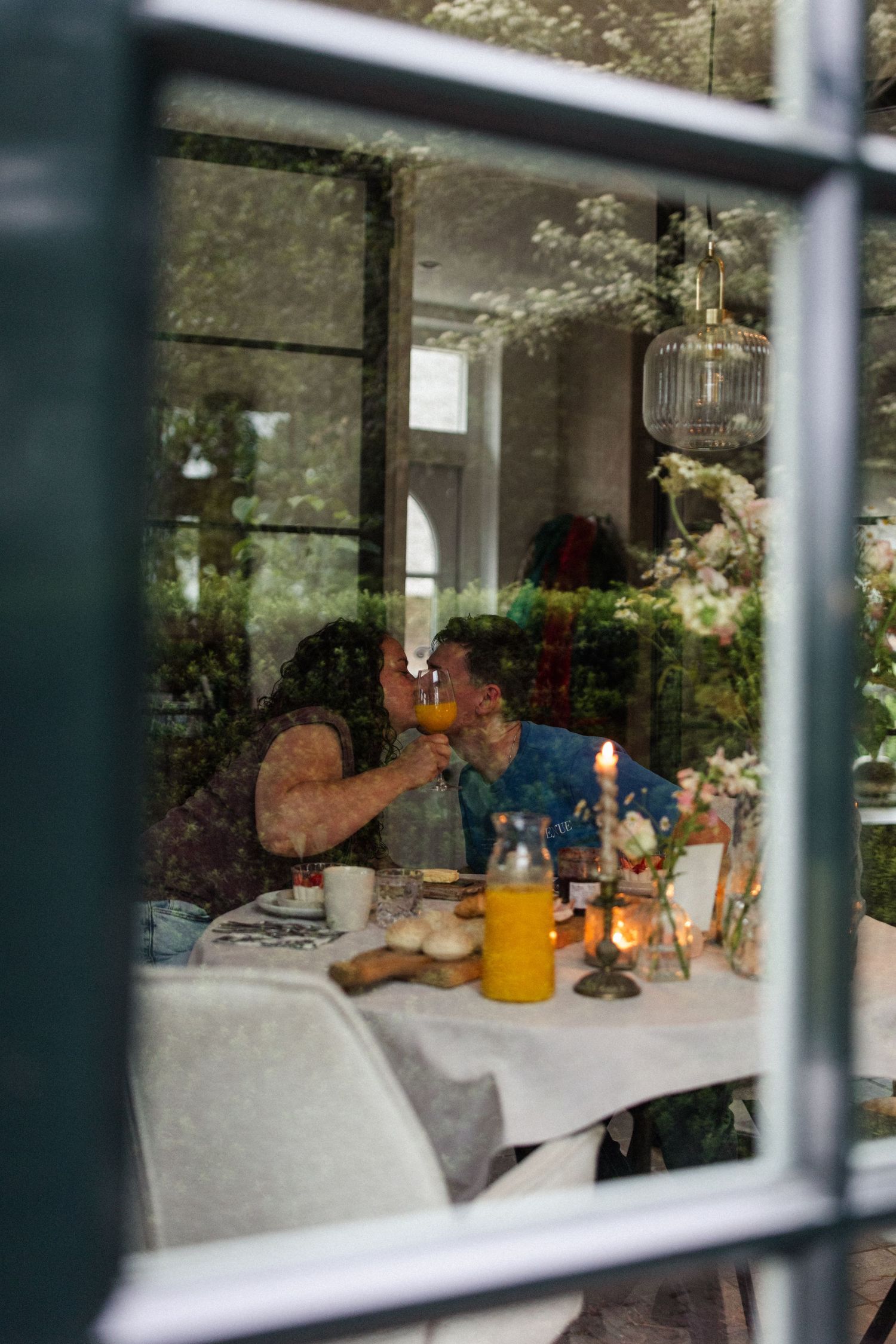 A candlelit dinner table setting viewed through a window pane with flowers and decorative elements.