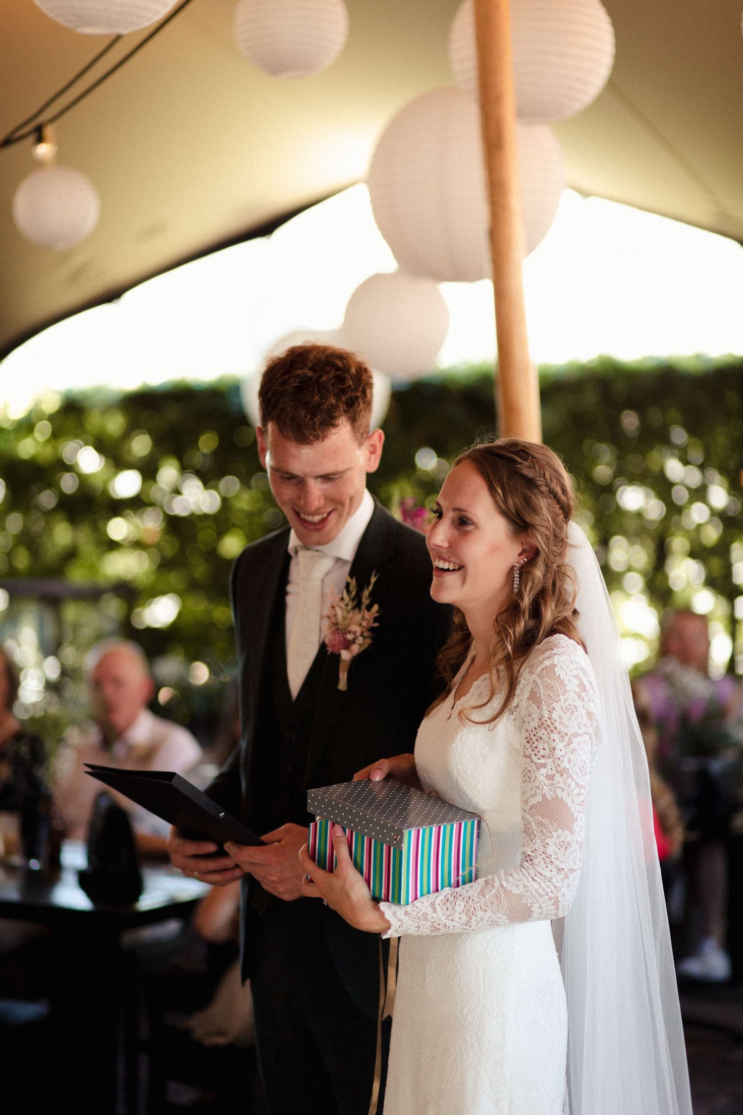 Newlyweds share a joyful moment while receiving a blue striped gift box under a tent with paper lantern decorations.