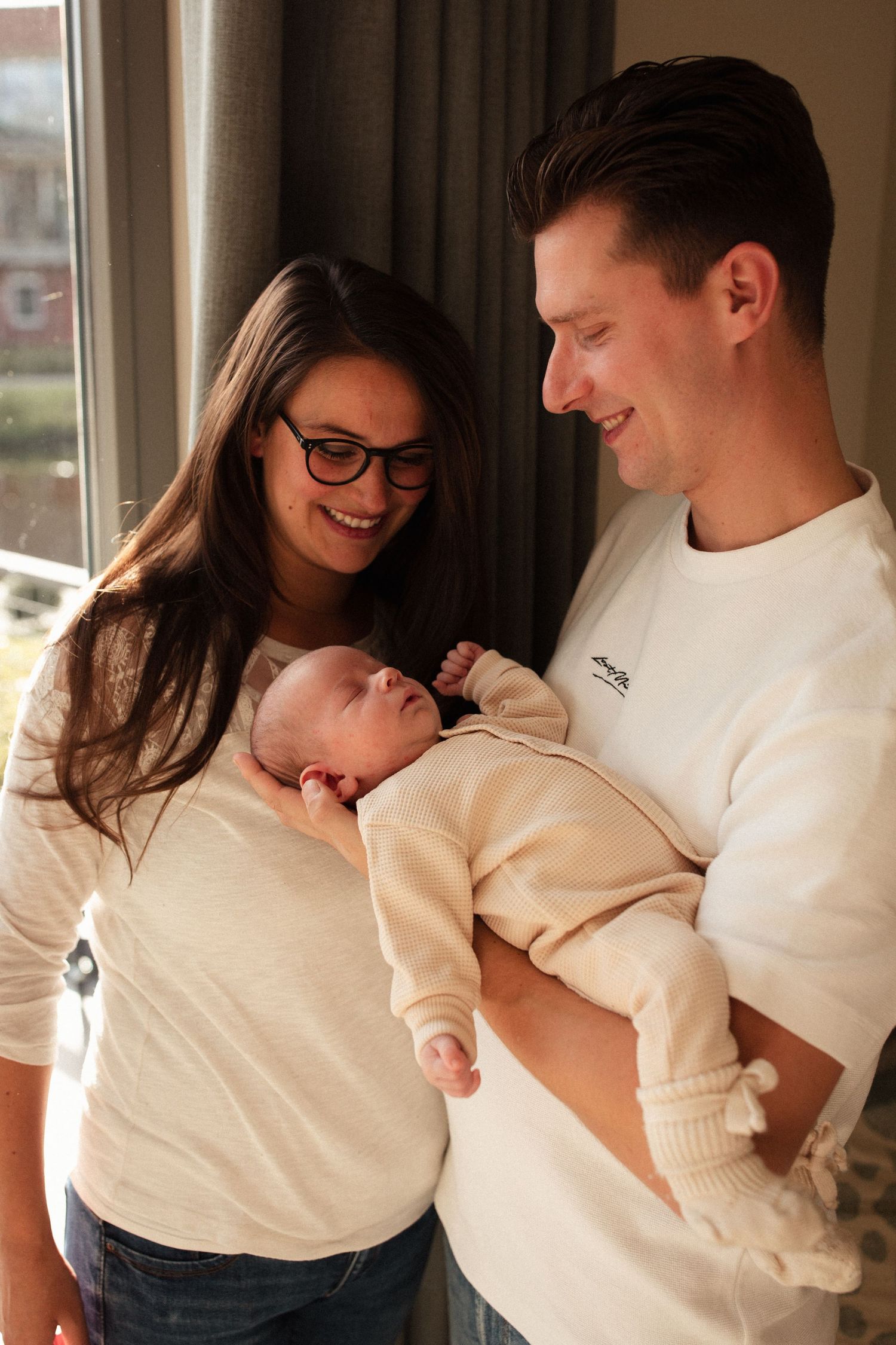 Candid family moment with newborn in cream colored clothing by window.