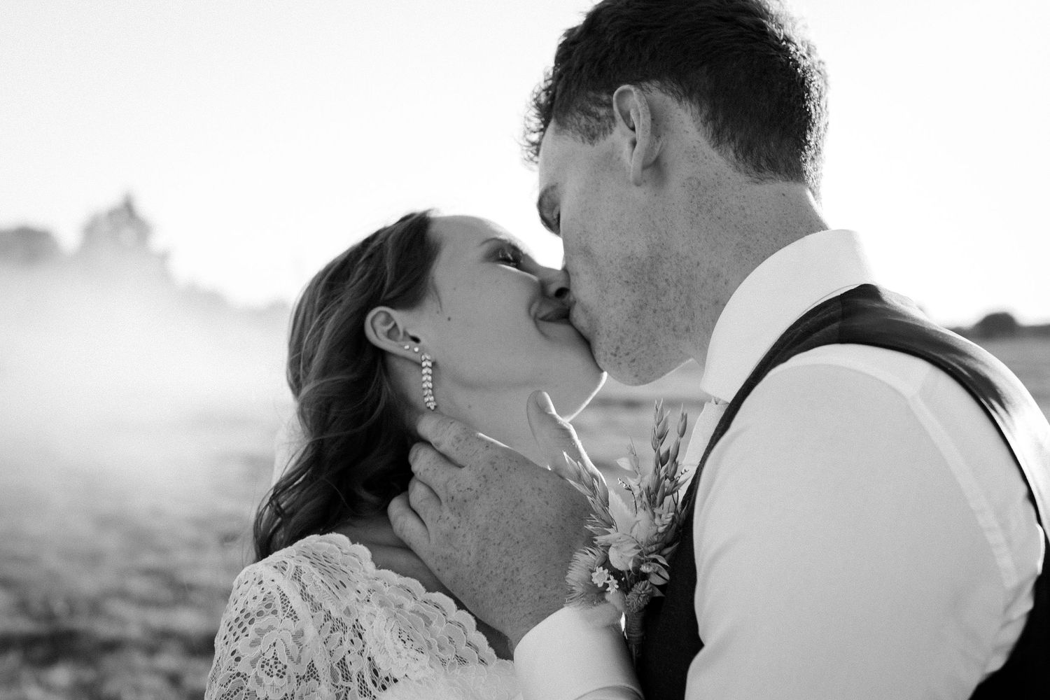 A romantic black and white wedding portrait captures a couple kissing in an ethereal misty outdoor setting.