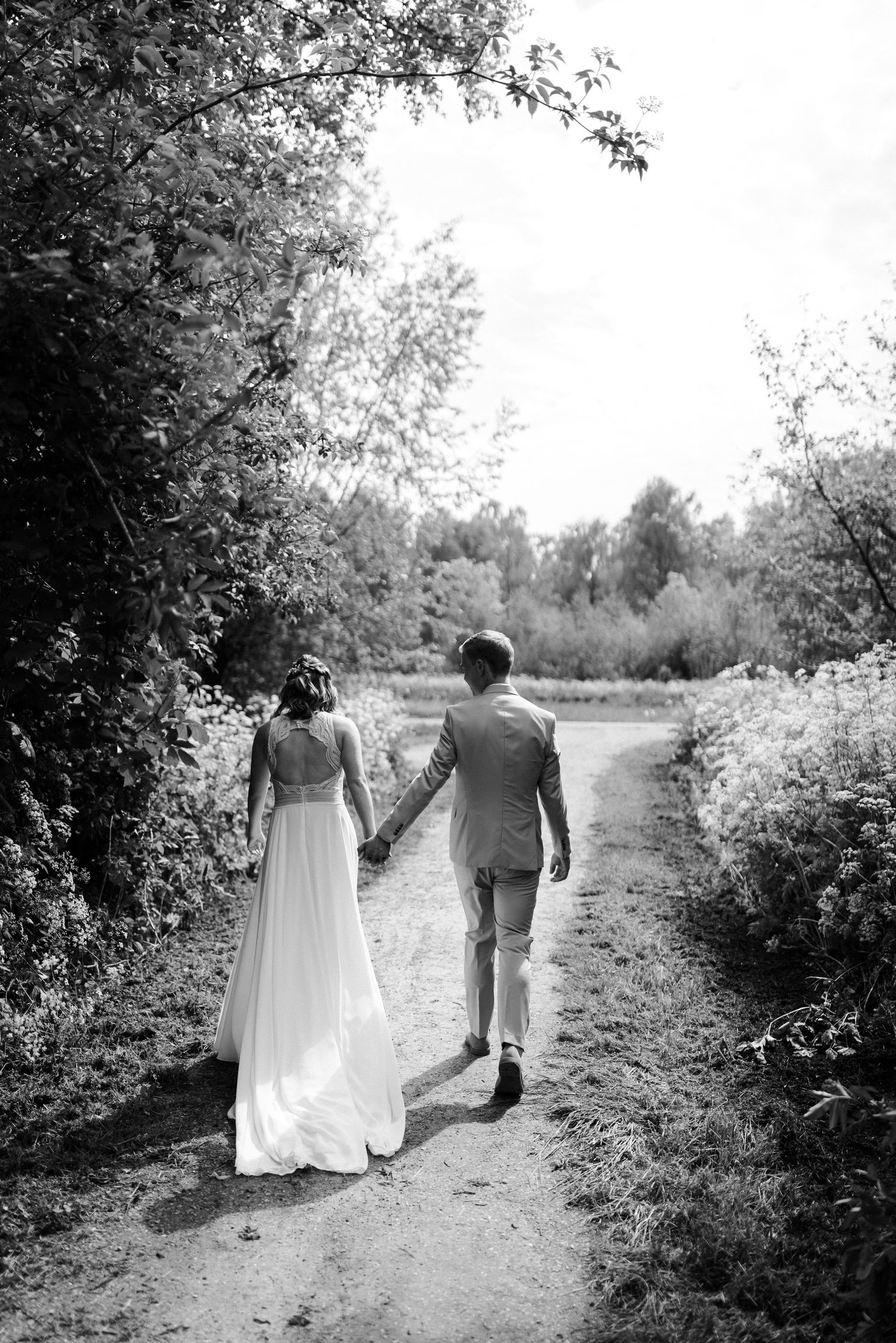 Black and white photo of a couple walking hand in hand down a nature path in wedding attire.