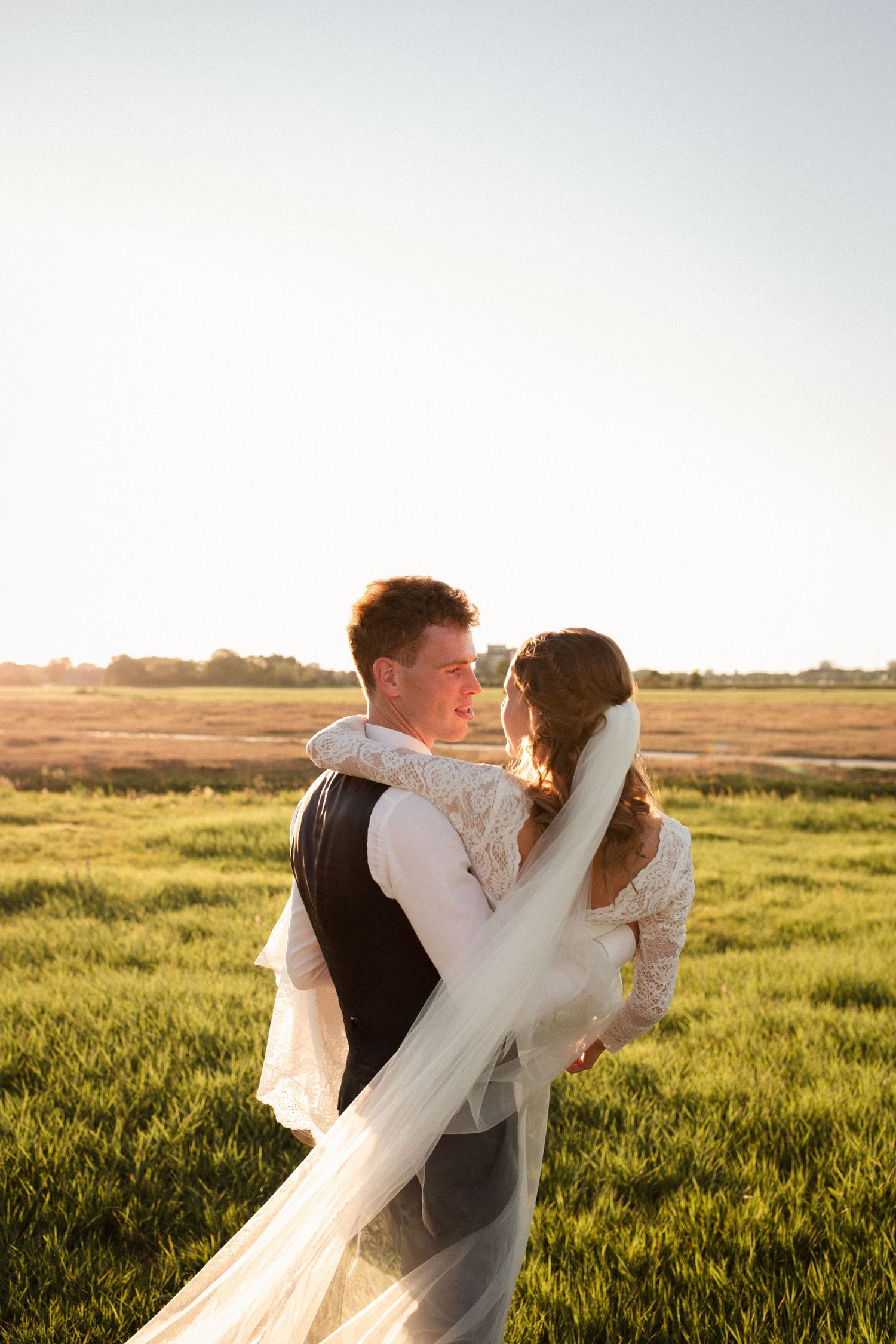 Couple embraces in golden sunlit field with flowing white veil during sunset wedding photoshoot.
