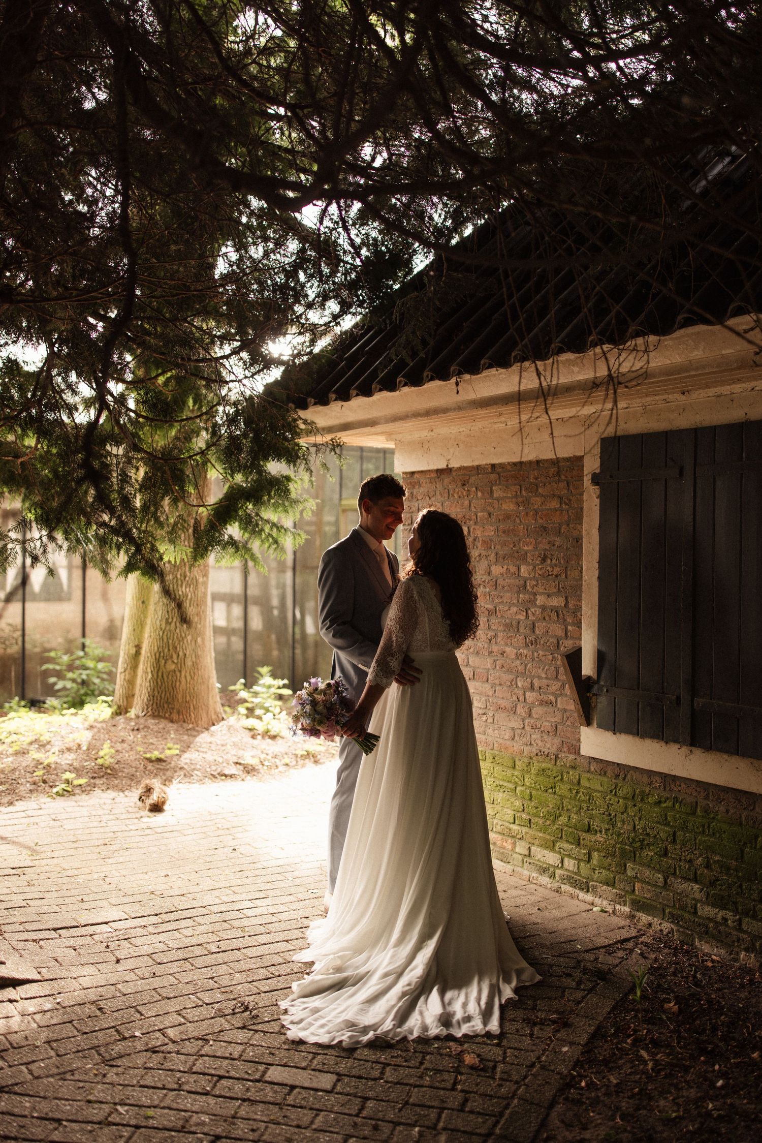 A romantic wedding couple embraces on a brick pathway under overhanging trees with dreamy backlit lighting.