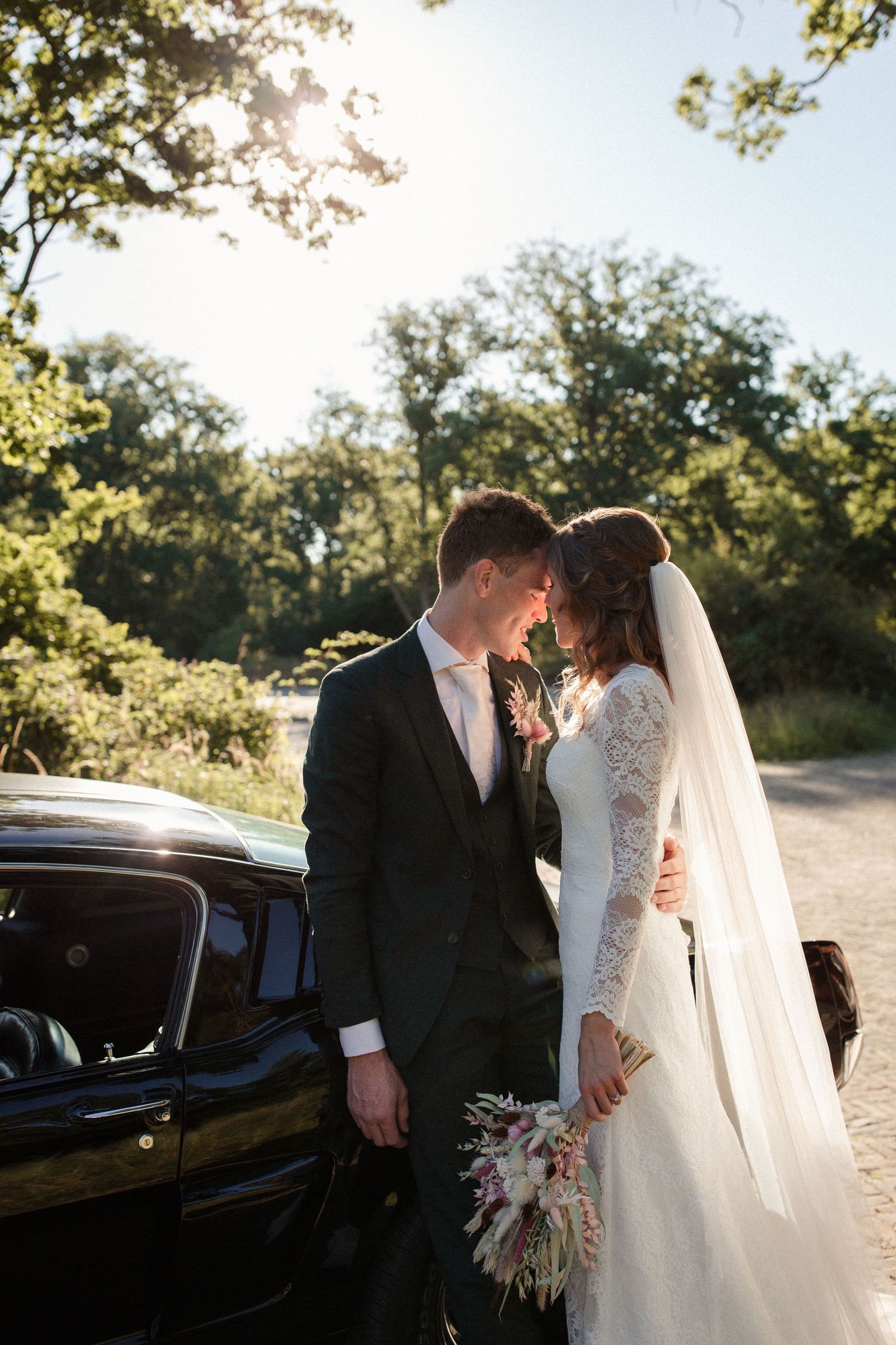 Two newlyweds share an intimate moment beside a vintage black car in golden sunlight during outdoor wedding shoot.