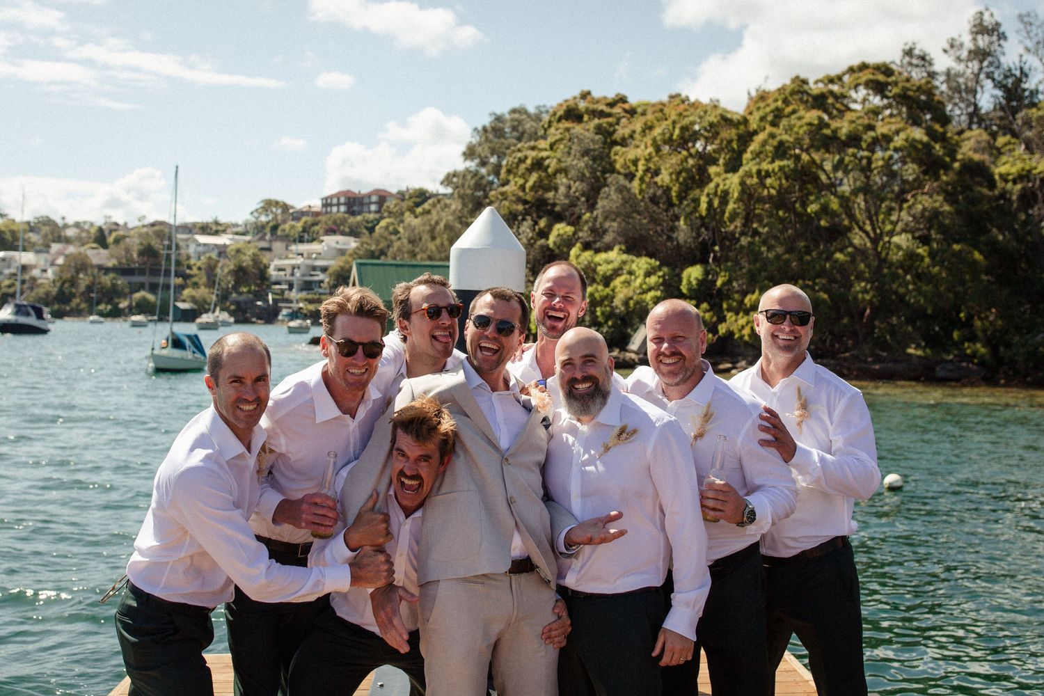 A cheerful wedding party in white attire poses together by the waterfront on a sunny day.