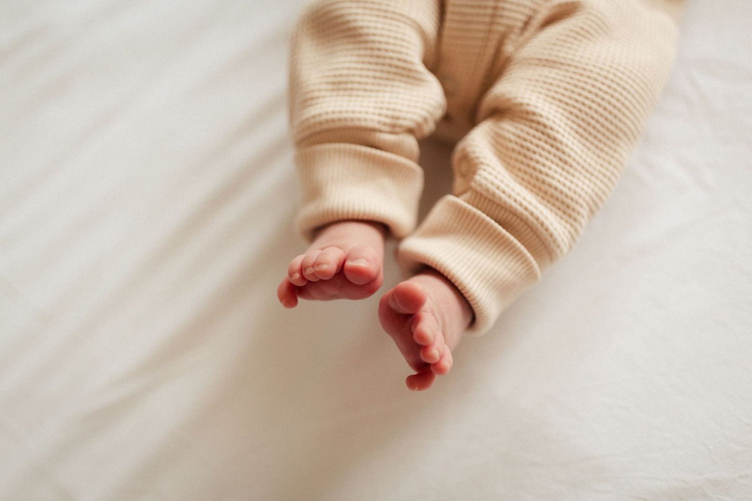 Tiny baby feet in beige knit pants against white fabric.
