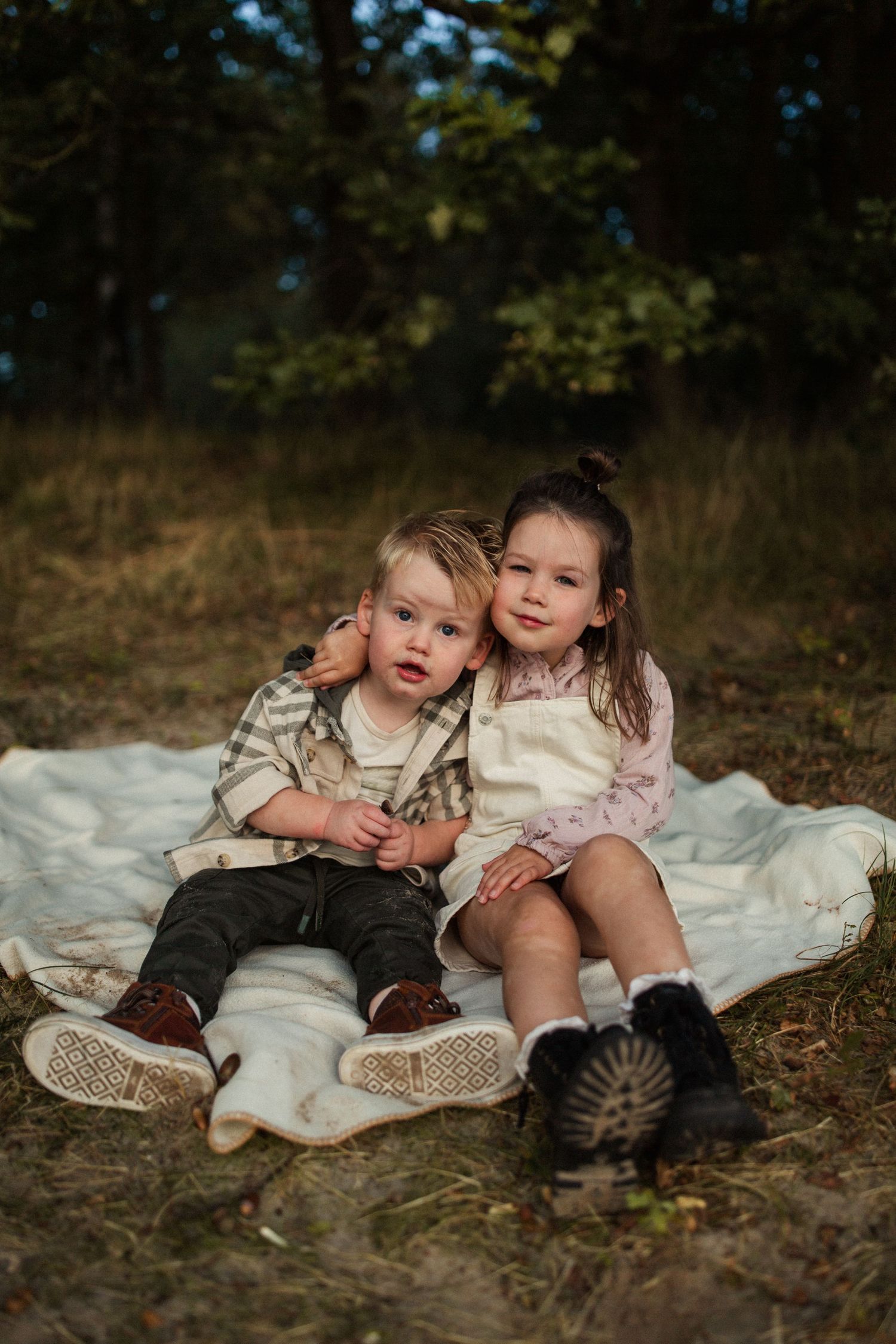 Two children sitting close together on a blanket in a wooded setting at golden hour.