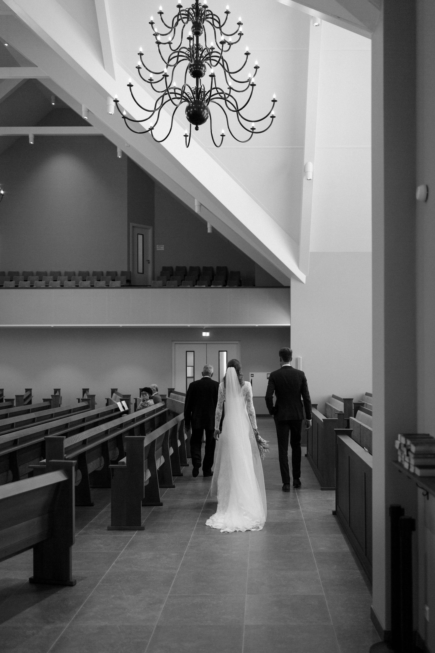 Black and white photo of a wedding party walking down a church aisle under an elegant chandelier.