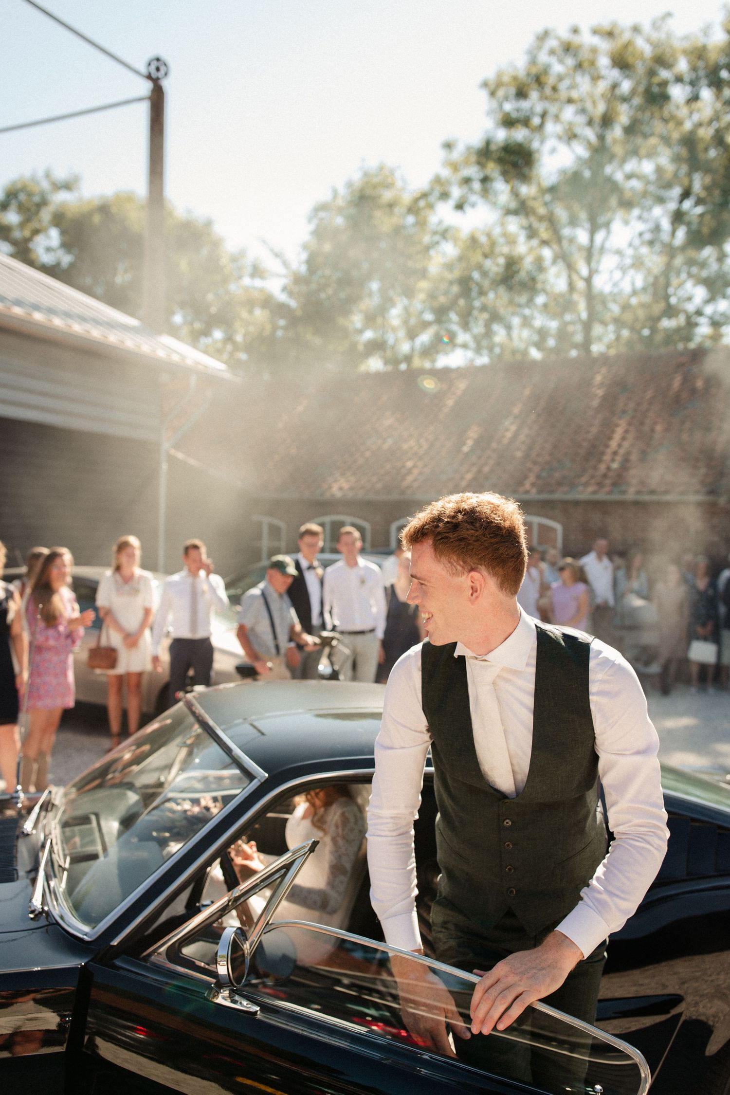 A person in formal attire stands beside a classic black car at an outdoor event on a sunny day with guests in background.