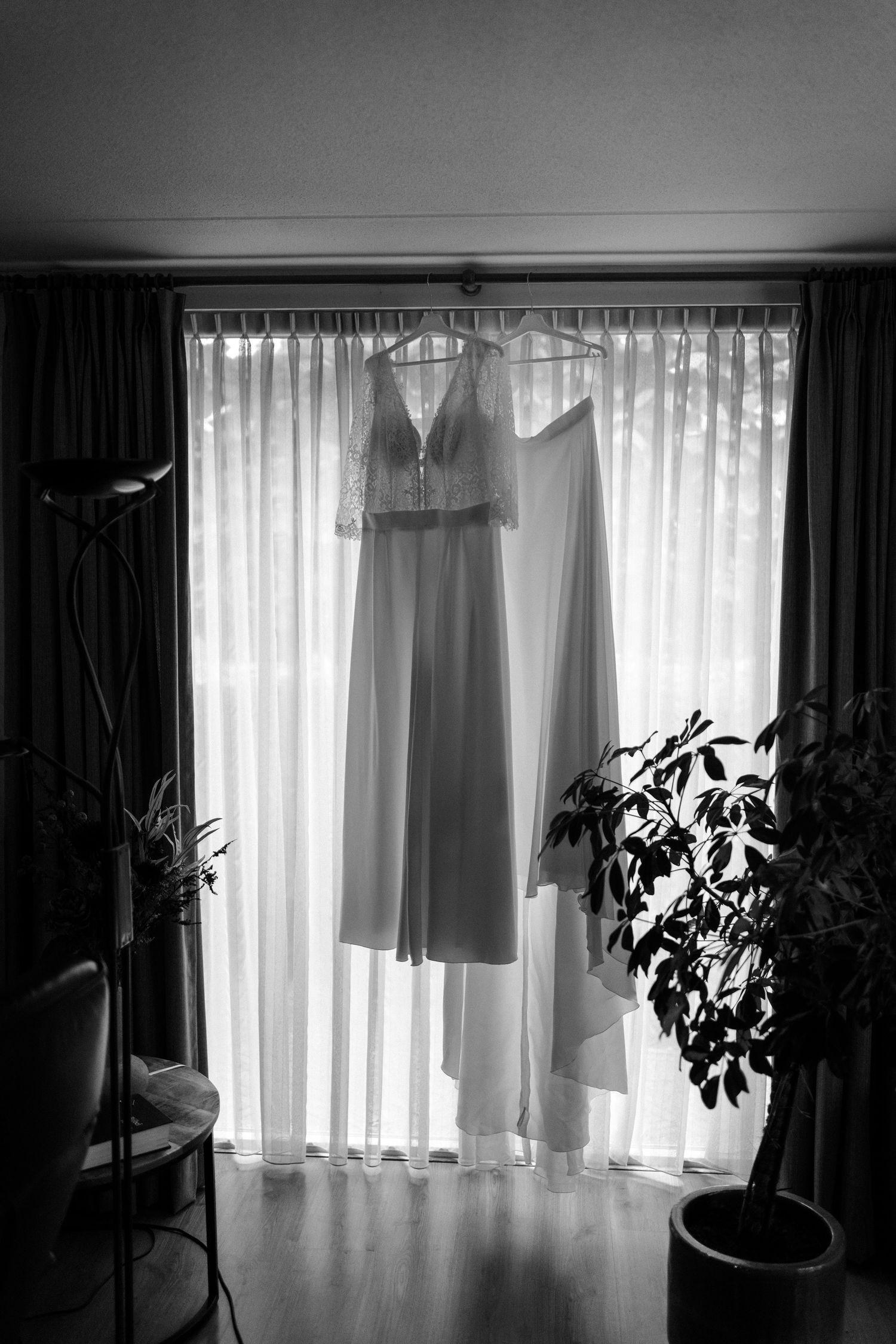 Two elegant wedding dresses hang in silhouette against a window with sheer curtains and fairy lights.